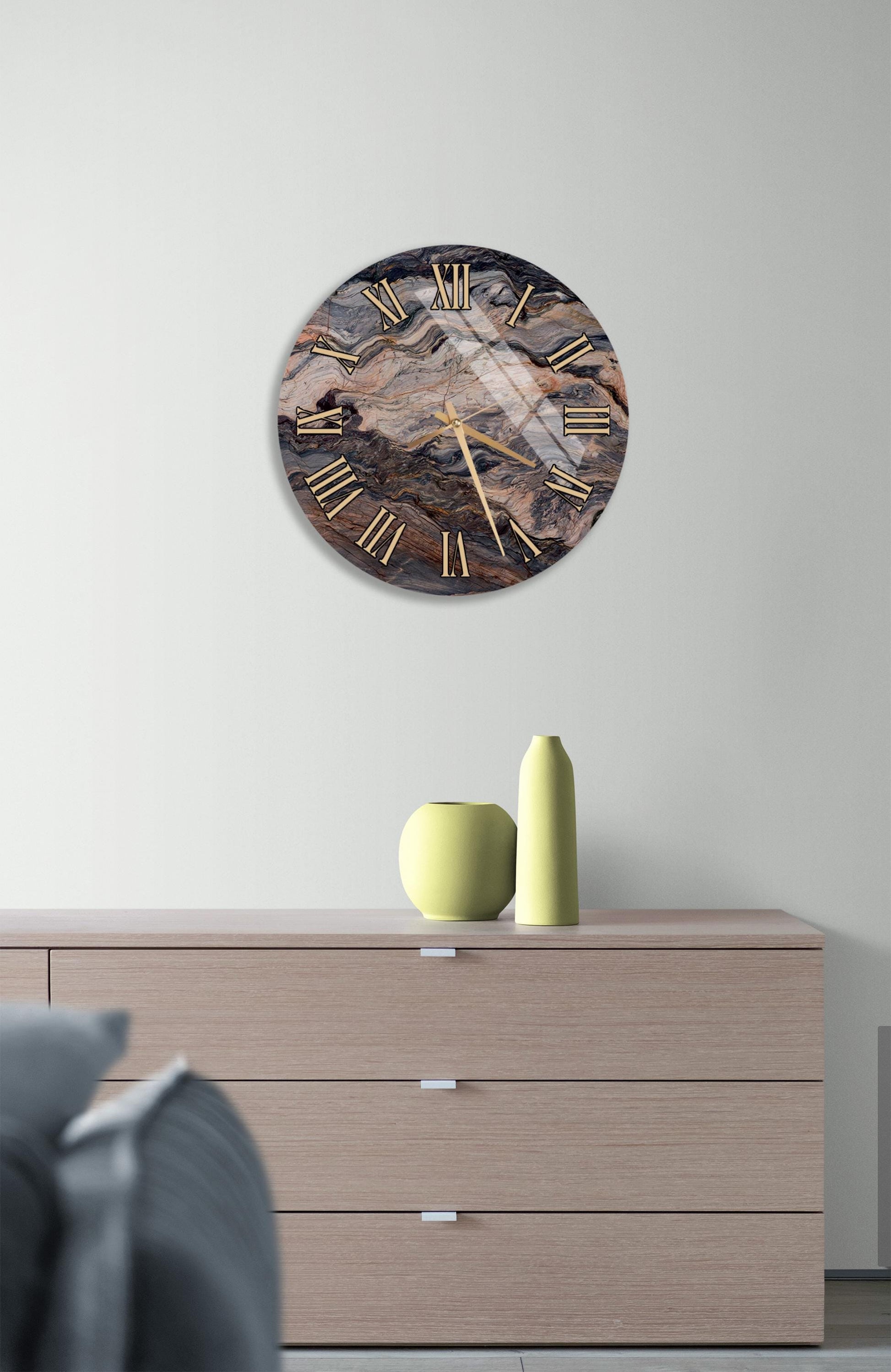 a modern interior design with a large round clock on a wall above a wooden dresser. The clock has a marble-like design and gold hands and numbers. The dresser has three drawers and a small yellow vase on top.