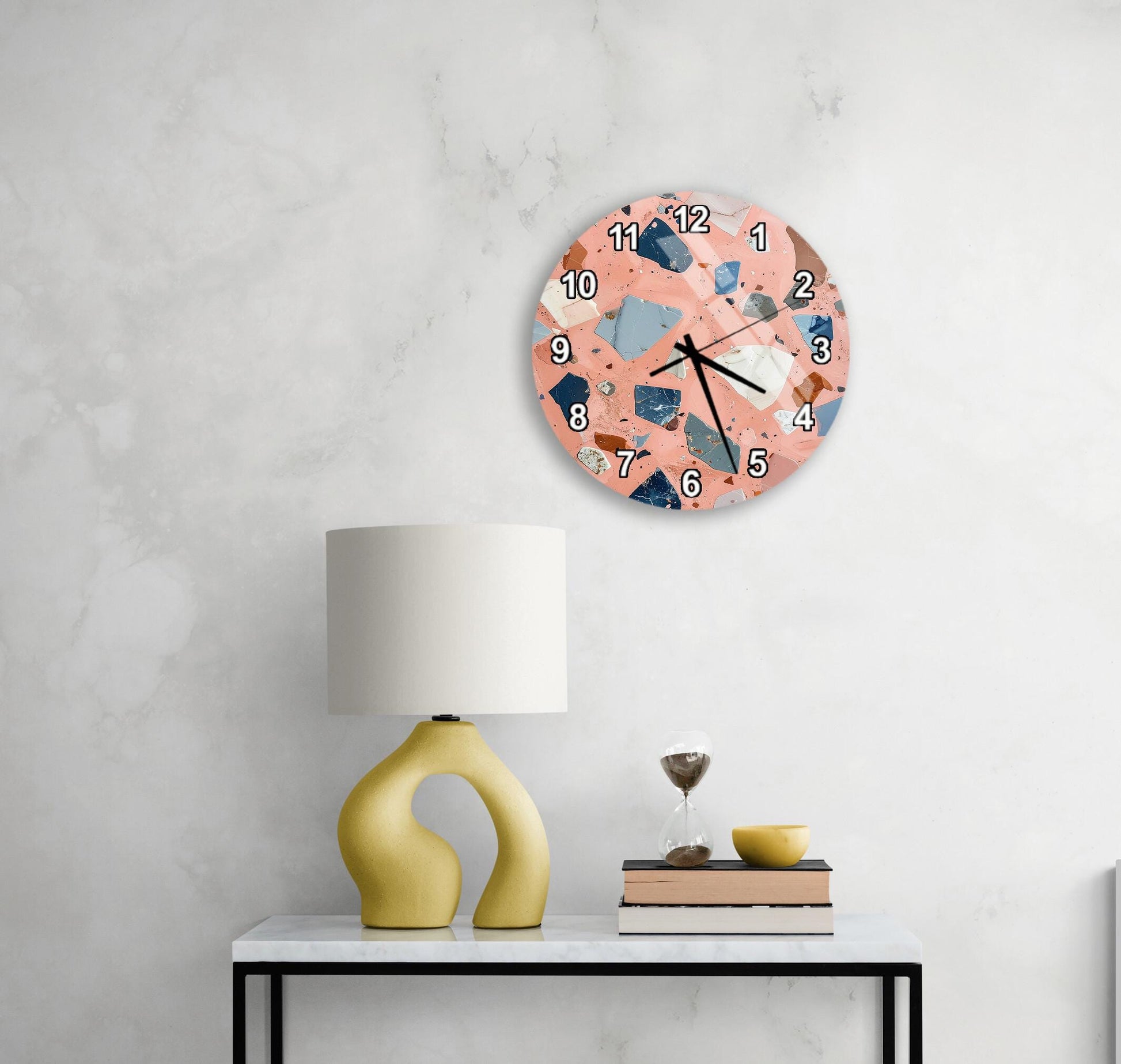 a modern, abstract-style clock with a pink and blue color scheme, mounted on a wall above a white table with a yellow lamp and a stack of books.