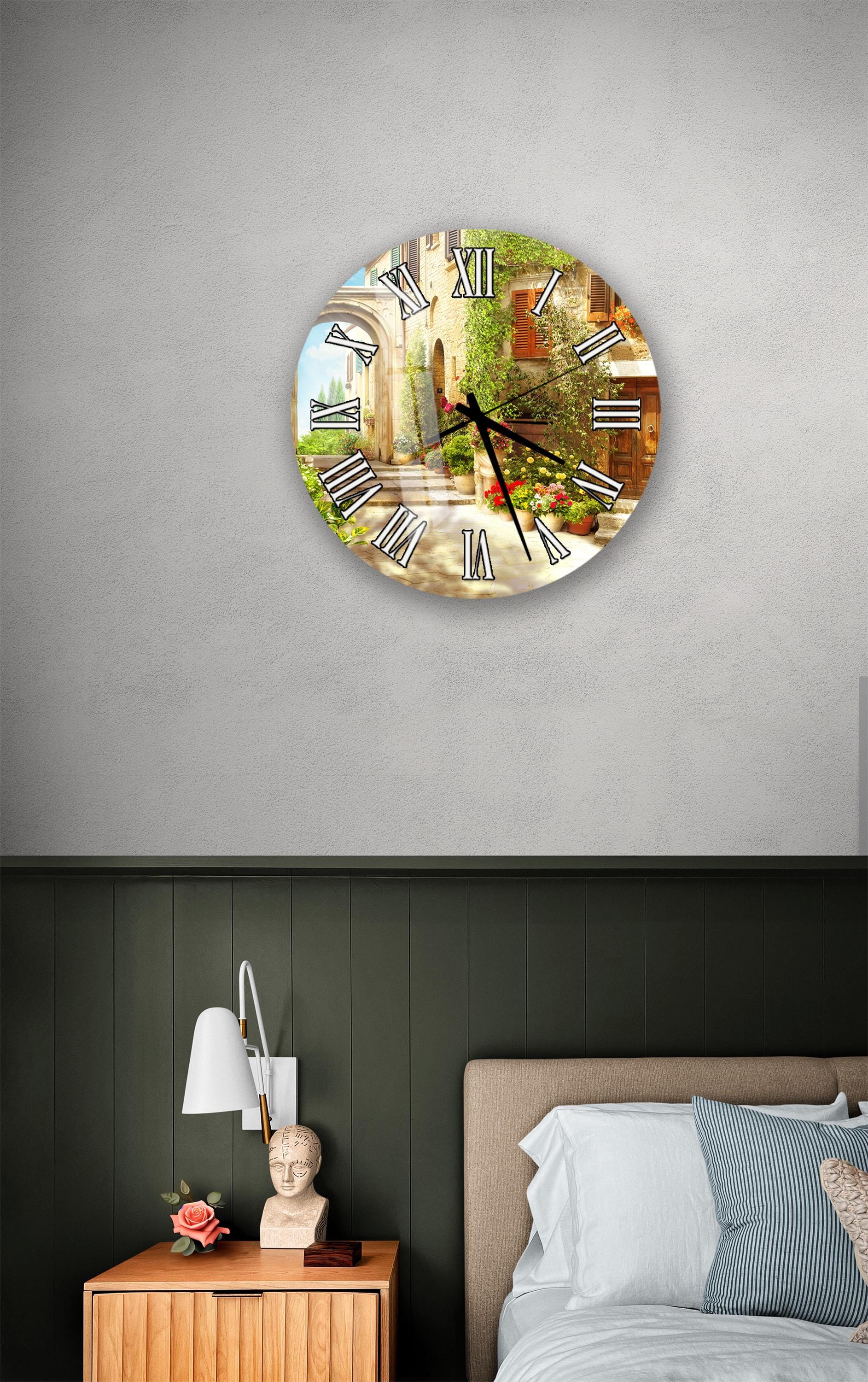 a wall clock with a scenic image of a building and a garden displayed on its face, mounted on a wall above a bedside table with a lamp and a small statue.