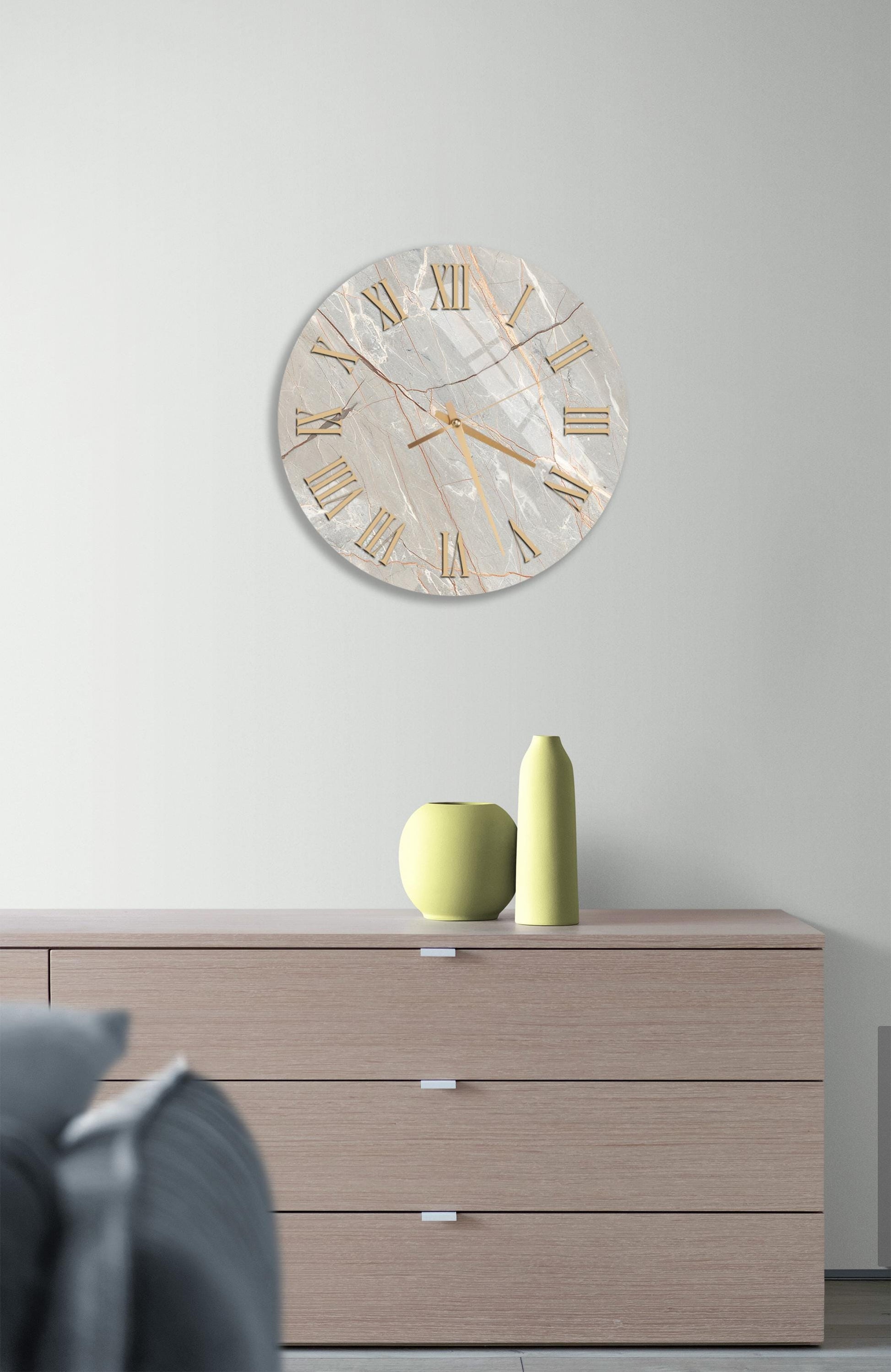 a modern living room with a large round clock on the wall above a wooden dresser. The clock has a marble-like finish and gold Roman numerals. On the dresser, there are two yellow vases and a small green vase, creating a stylish and cohesive decor.