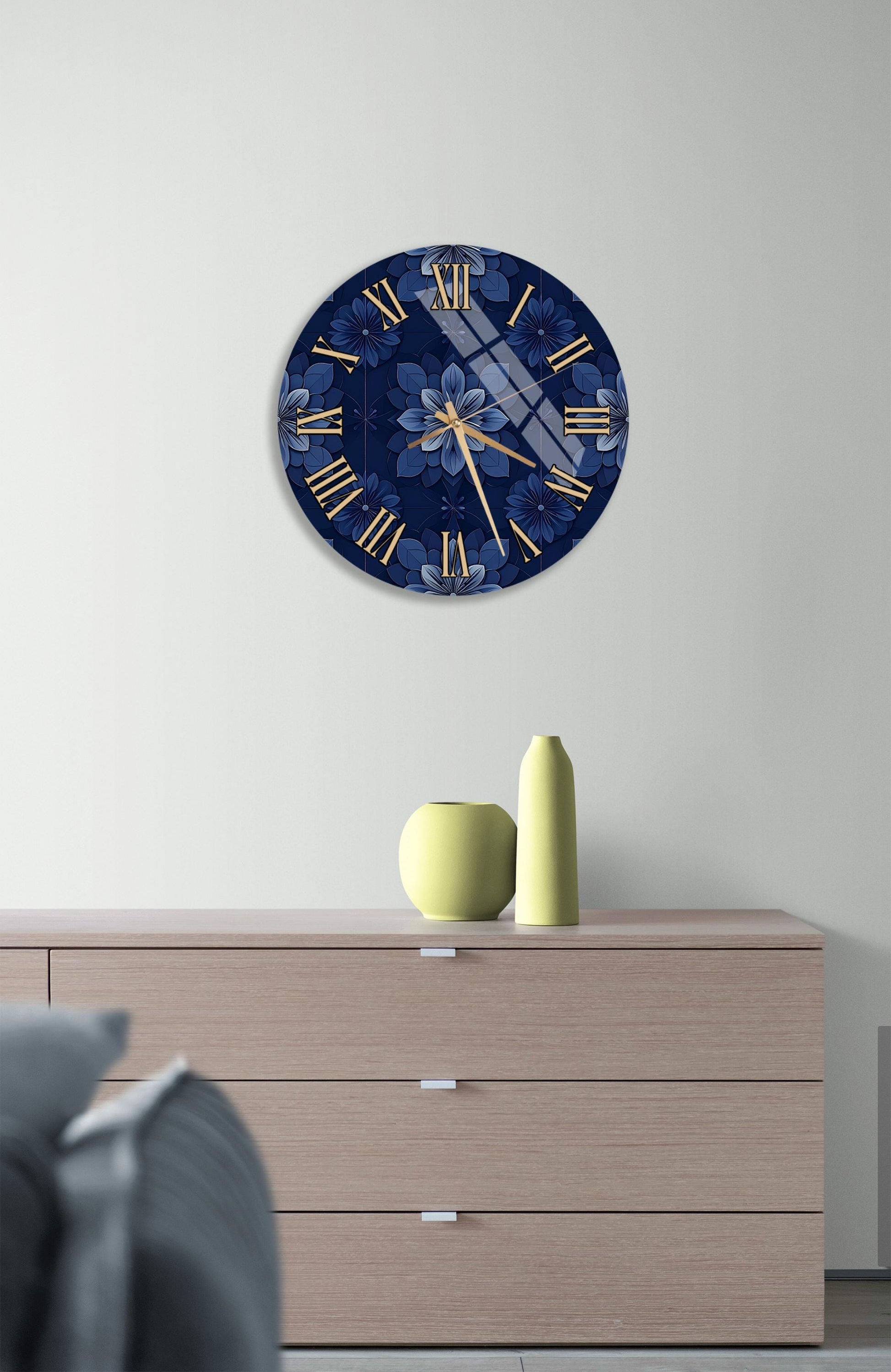 a wall clock with a blue floral design, mounted on a wall above a wooden dresser. The dresser has a vase and a small yellow object on top of it.