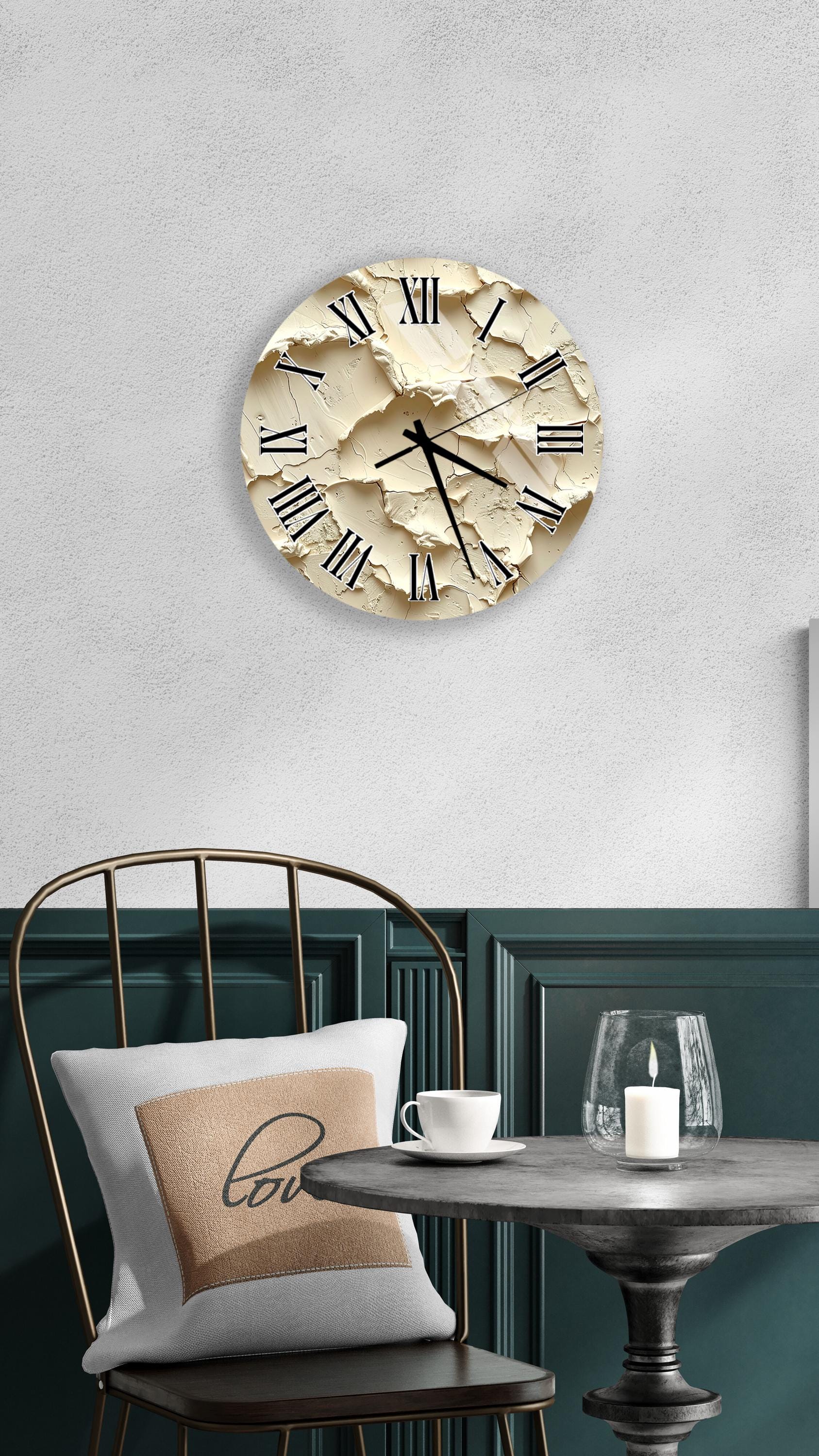 a round clock with a beige face and black Roman numerals mounted on a white wall above a black metal chair with a white pillow. The chair is positioned in front of a small table with a glass and a candle on it.