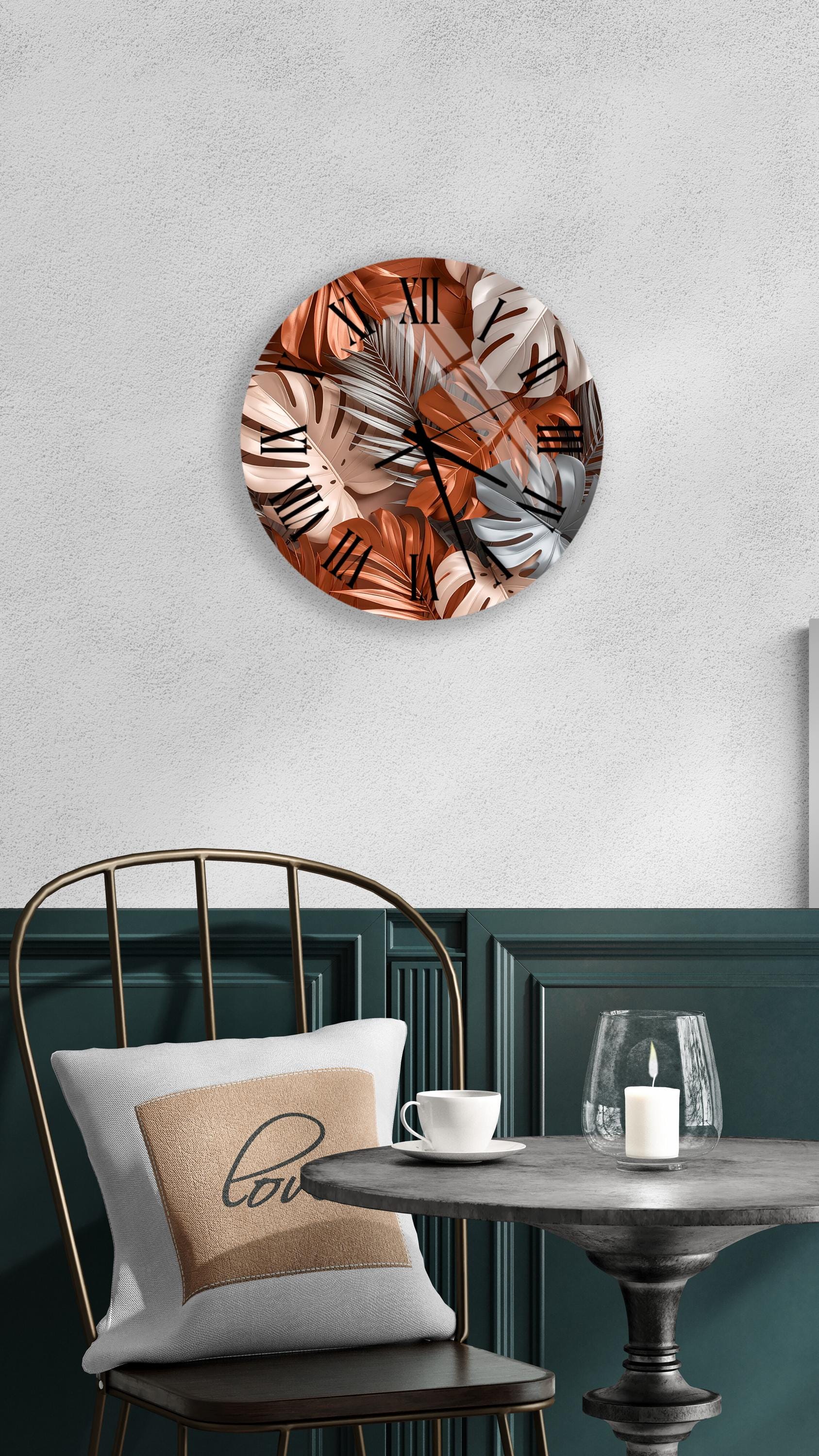 a round wall clock with a tropical leaf pattern, a white table with a candle and a cup, and a green chair with a pillow.