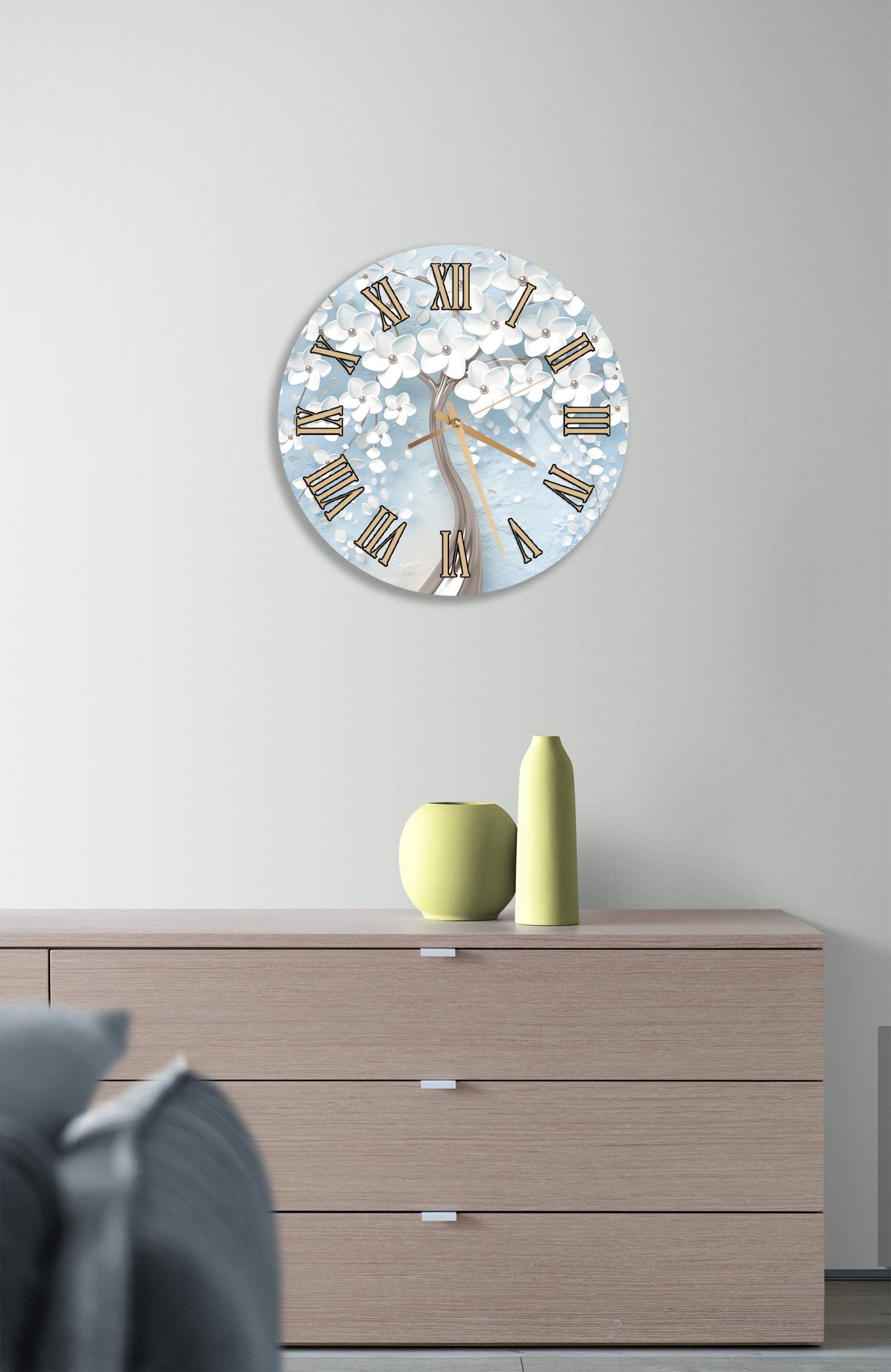 a wall clock with a floral design, mounted on a wall above a wooden dresser with a vase and a small sculpture on top.