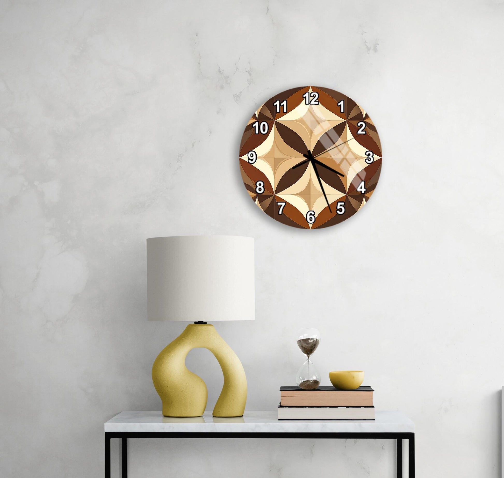 a modern wall clock with a brown and beige design, mounted on a white wall above a white table with a yellow lamp and a stack of books.