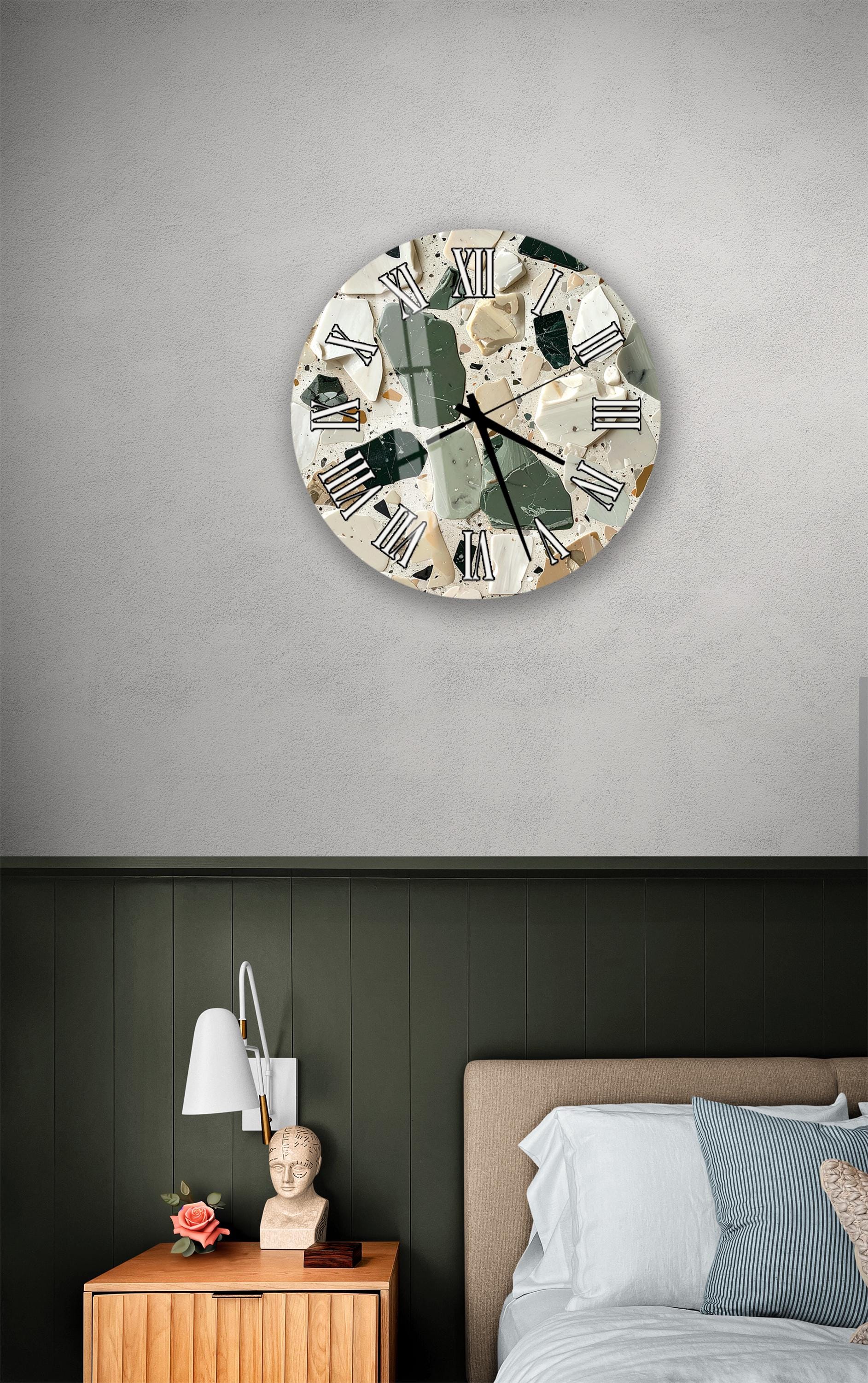 a bedroom setting with a round clock on the wall above a bed. The clock has a unique, mosaic-like design with various colors and patterns. The room has a green wall, a wooden nightstand, and a lamp.