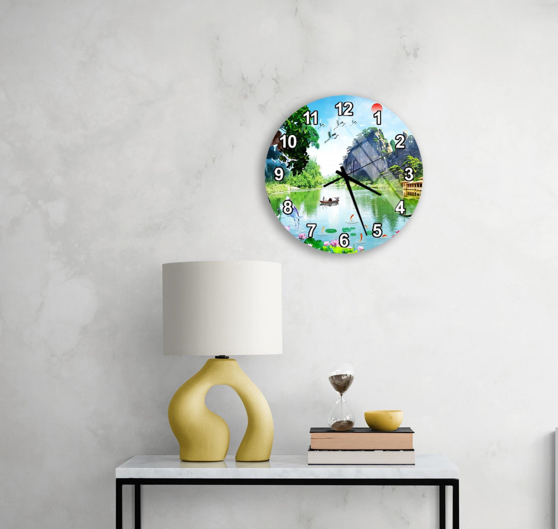 a round clock with a scenic image of a lake and trees on its face, mounted on a wall above a white table with a yellow lamp and a stack of books.