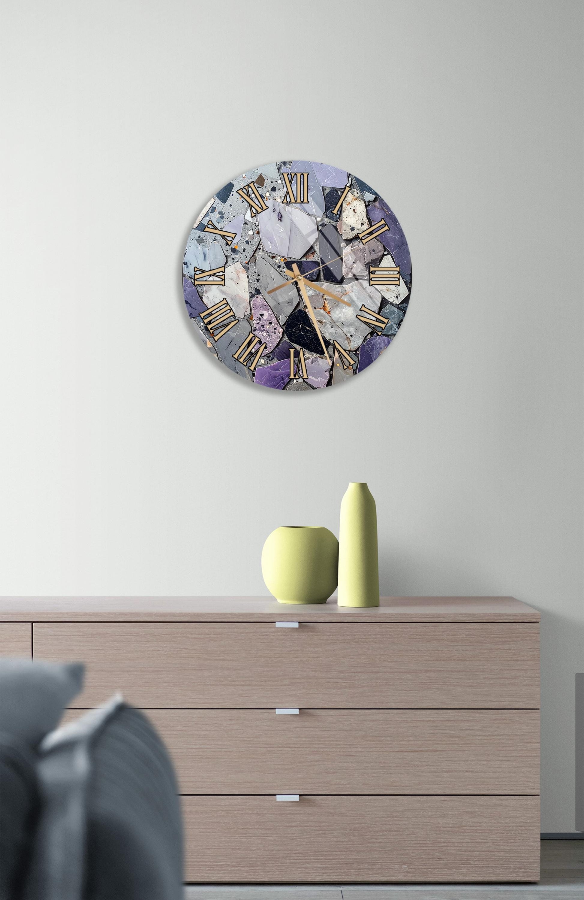 a modern clock with a mosaic design on the wall above a wooden dresser with three drawers. The clock has a combination of purple and gray colors, and the dresser has a minimalist design with a single yellow vase on top.