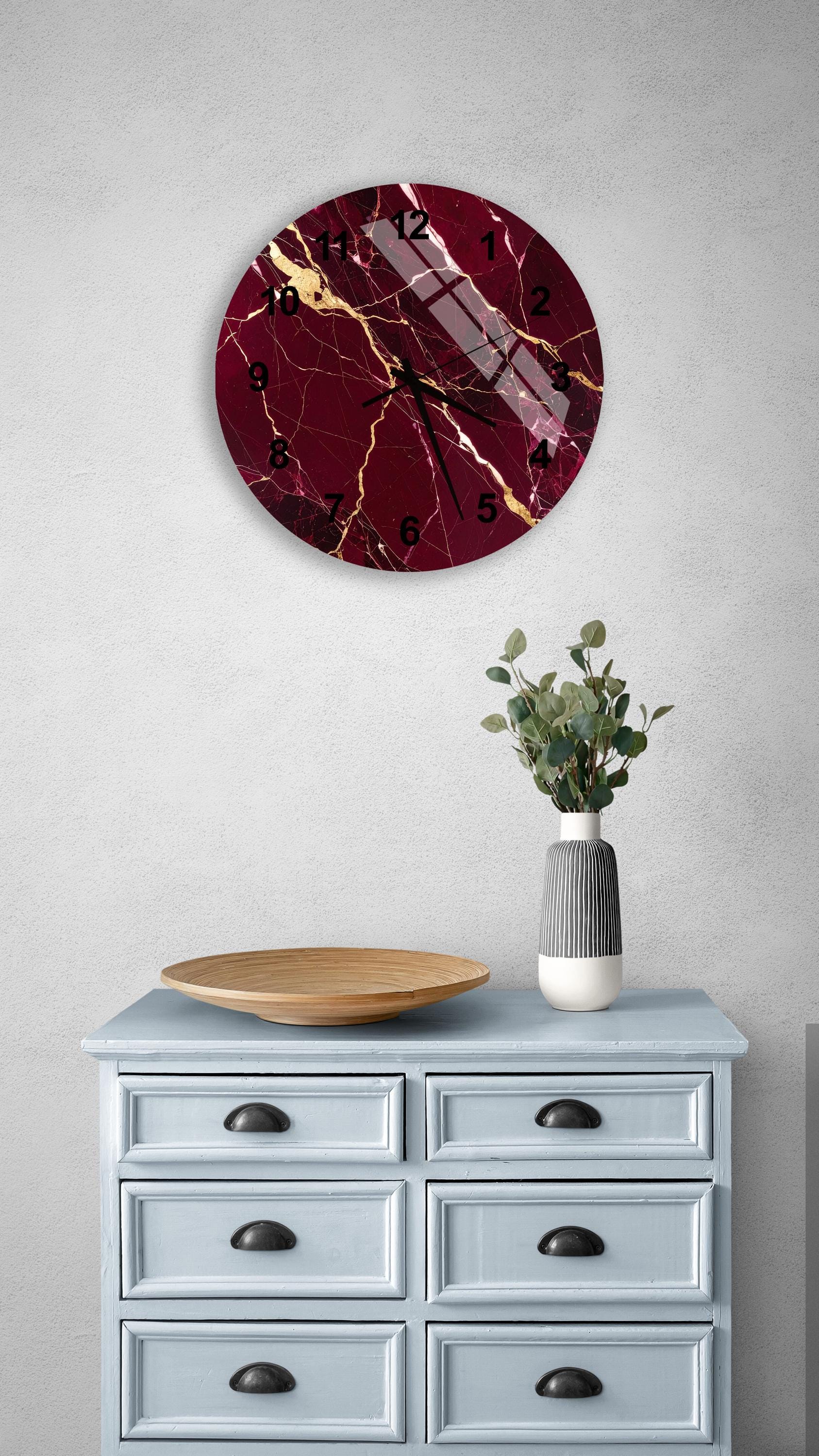 a wall clock with a maroon and gold design, mounted on a white wall above a light blue dresser with six drawers. The dresser also has a wooden tray and a vase with greenery on it.