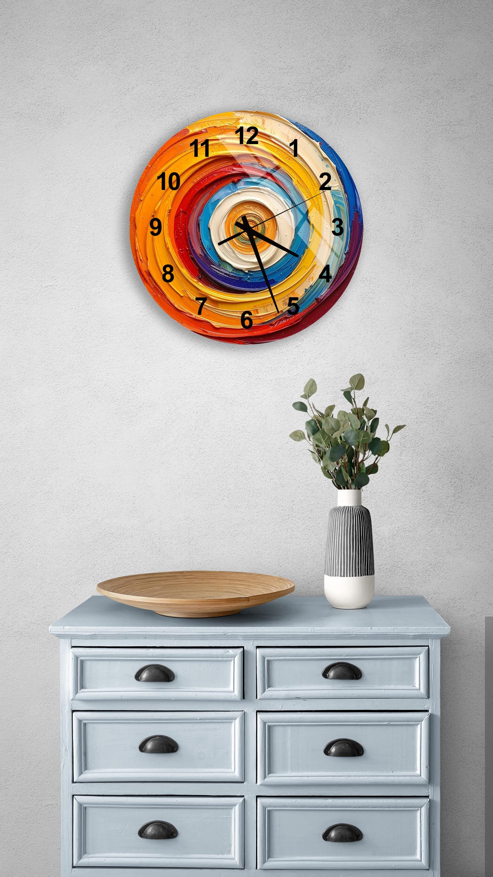 a wall clock with a colorful spiral design, mounted on a wall above a light blue dresser with six drawers. The dresser also has a vase with greenery and a wooden tray on top.