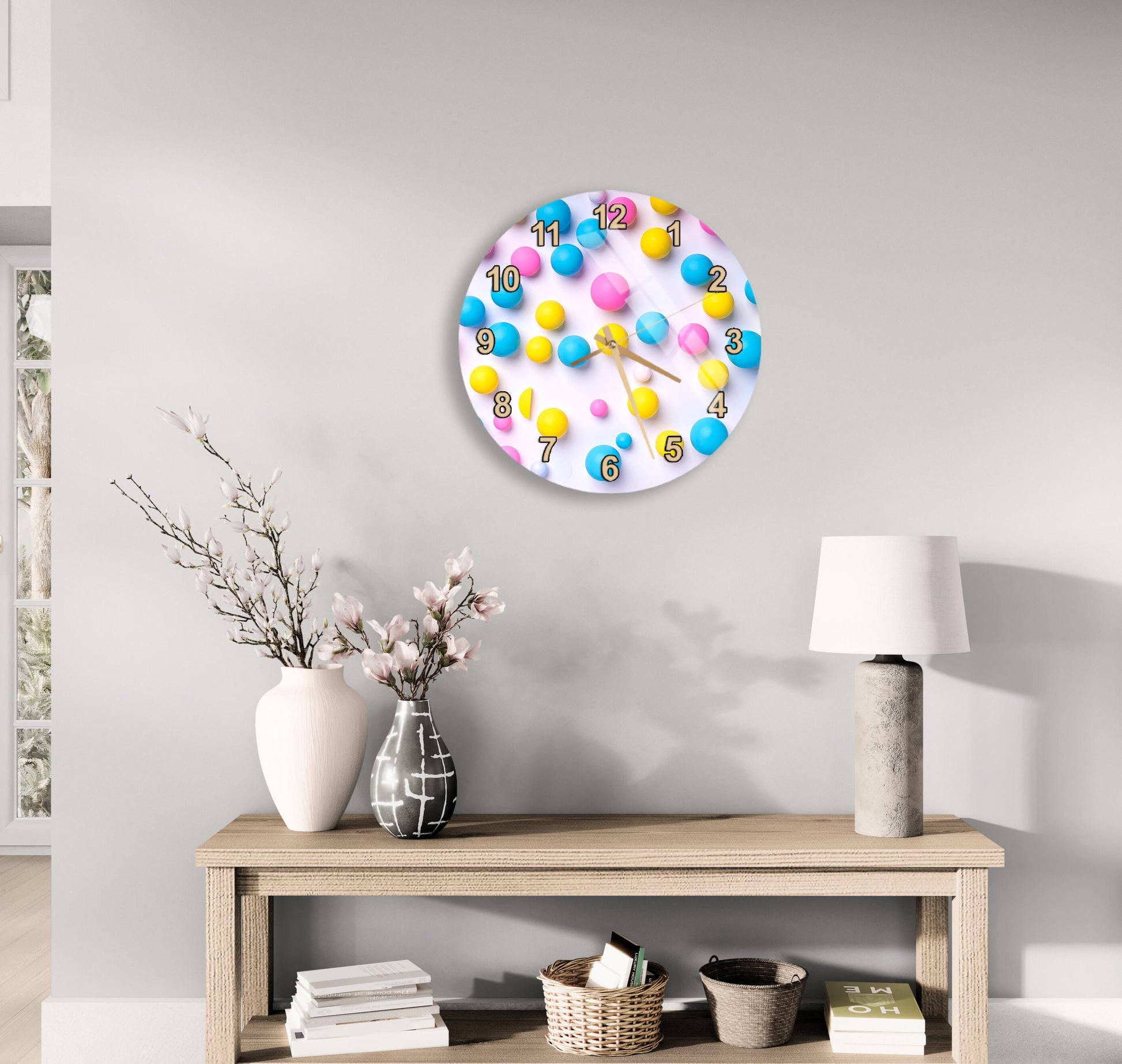 a clock with colorful balls on it, hanging on a wall above a wooden table with various decorative items such as a vase, a lamp, and some books.