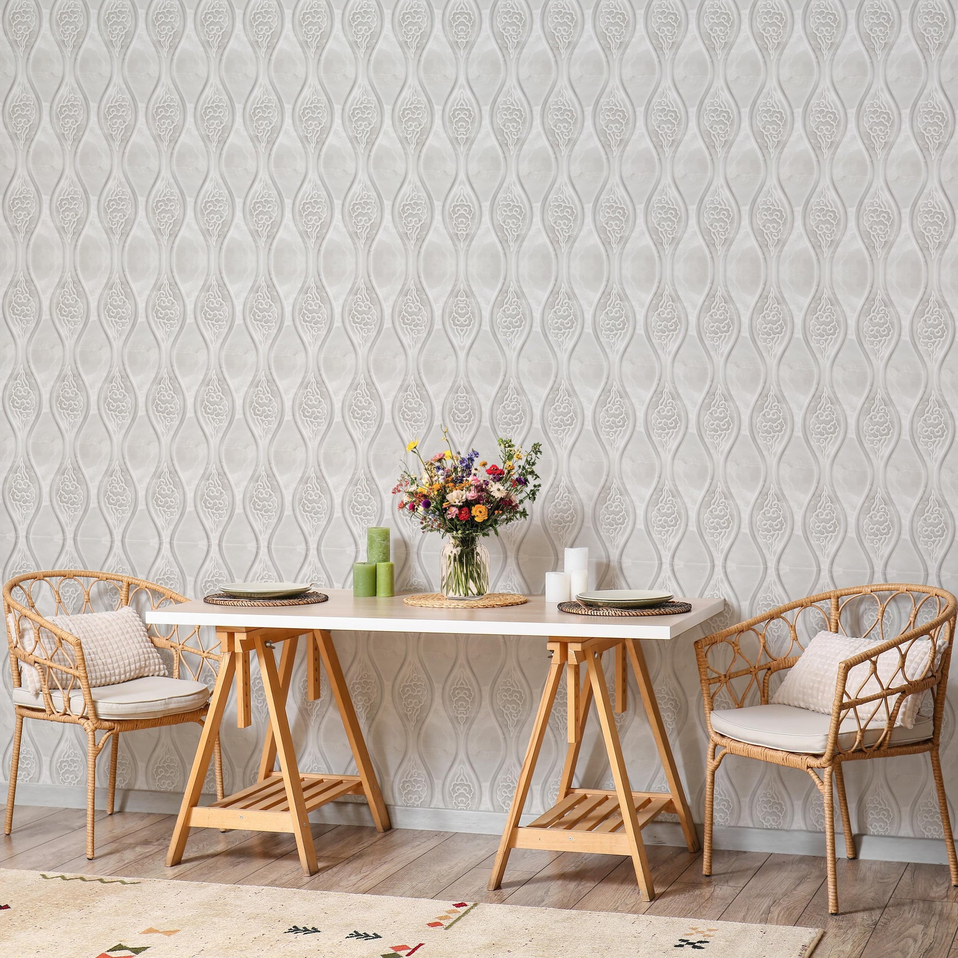a dining area with a white table, two chairs, and a vase of flowers. The chairs are made of wicker and have cushions. The table is set with plates and candles, creating a cozy and inviting atmosphere. The background has a patterned wallpaper, adding visual interest to the space.