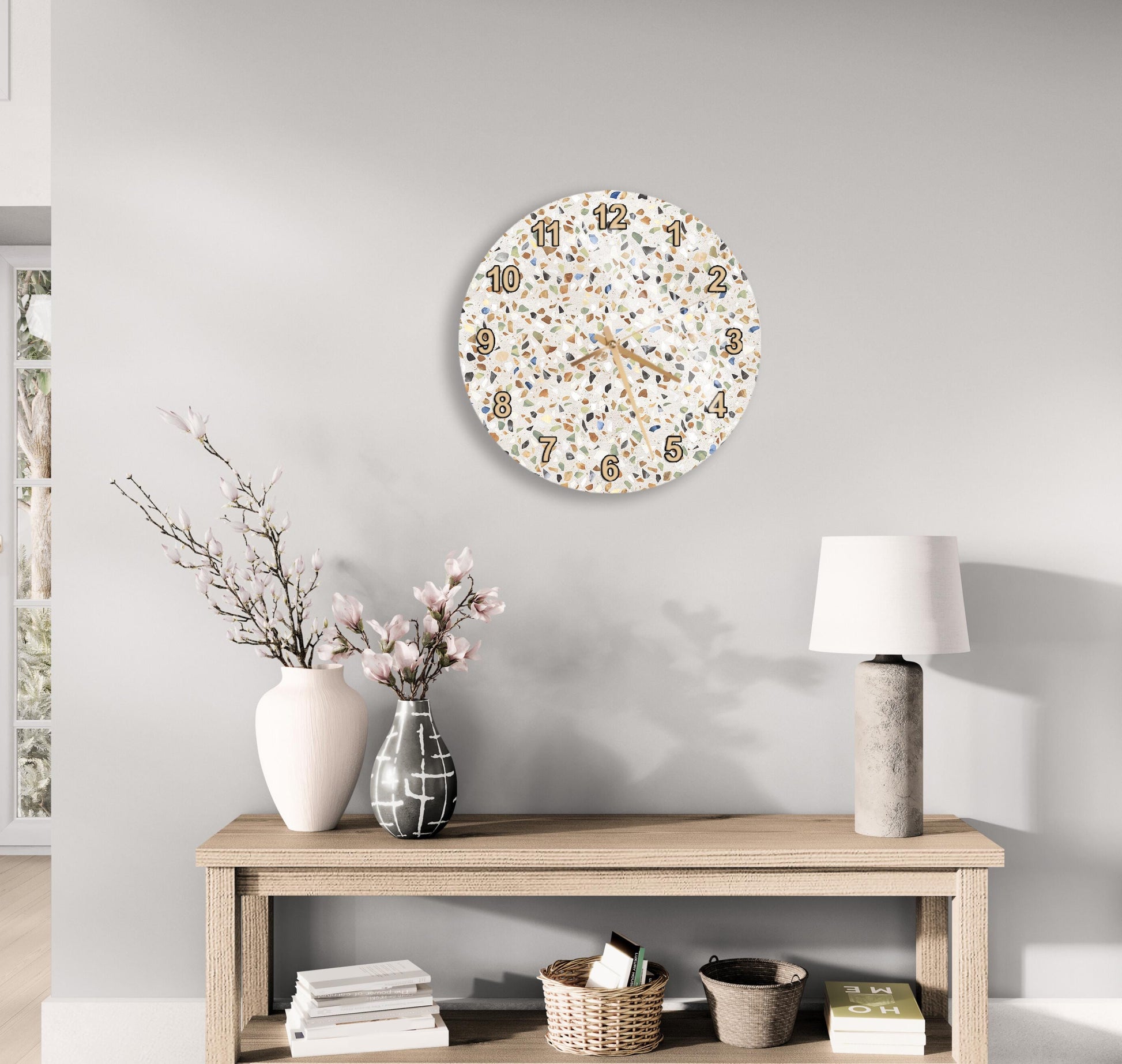 a modern and minimalist living room with a wooden console table, a round wall clock with a speckled design, a vase with pink flowers, and a lamp on a side table.