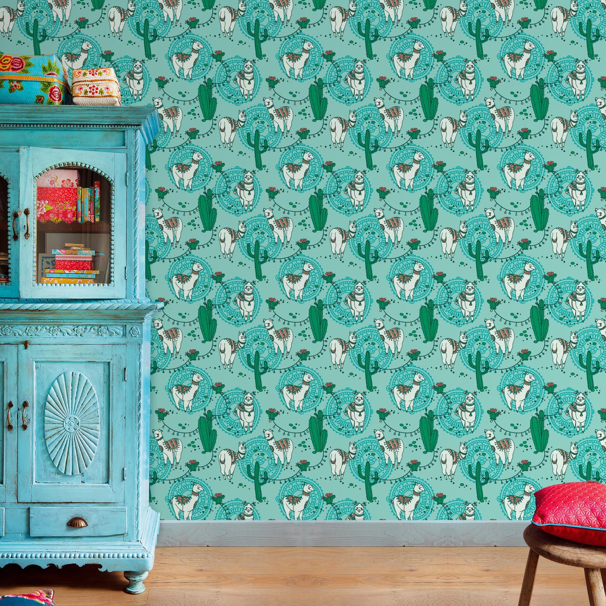 a vibrant turquoise wall with a repeating pattern of cacti and llamas, creating a lively and colorful atmosphere. A blue wooden cabinet with a decorative design is situated next to the wall, and a red cushion is placed on a wooden stool in the foreground.