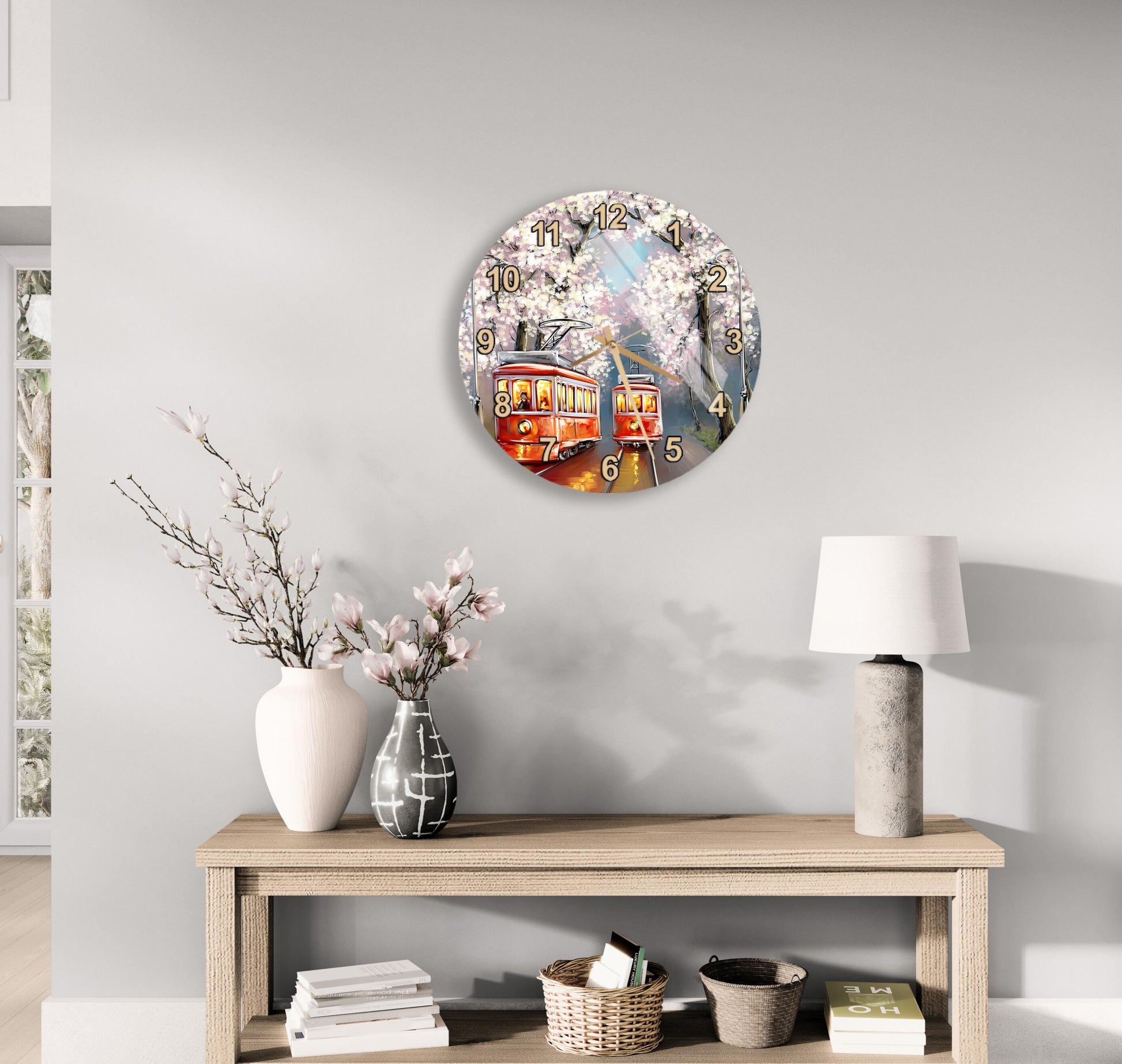 a round clock with a floral design hanging on a wall above a wooden table with various decorative items, including a vase with flowers, a lamp, and a stack of books.