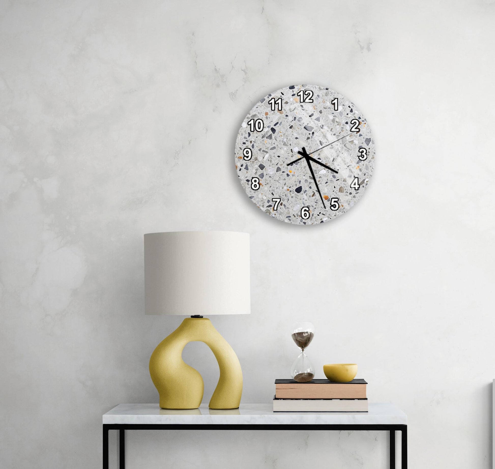 a modern living room with a white marble wall, a round wall clock with a speckled design, a yellow lamp with a unique curved base, and a small table with a stack of books and a glass of water.