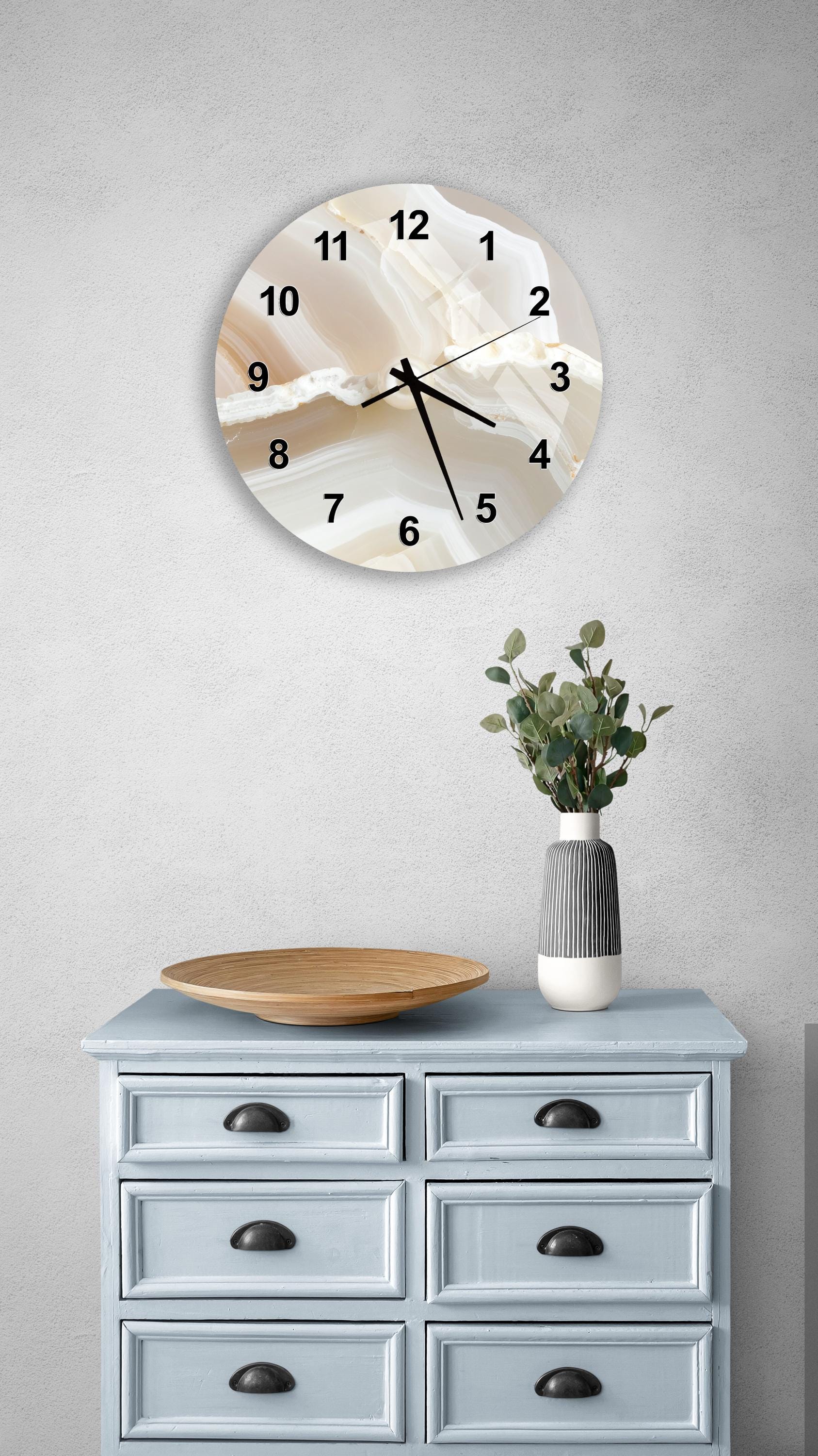a wall clock with a marble-like design, a wooden bowl, and a vase with greenery on a light blue dresser.