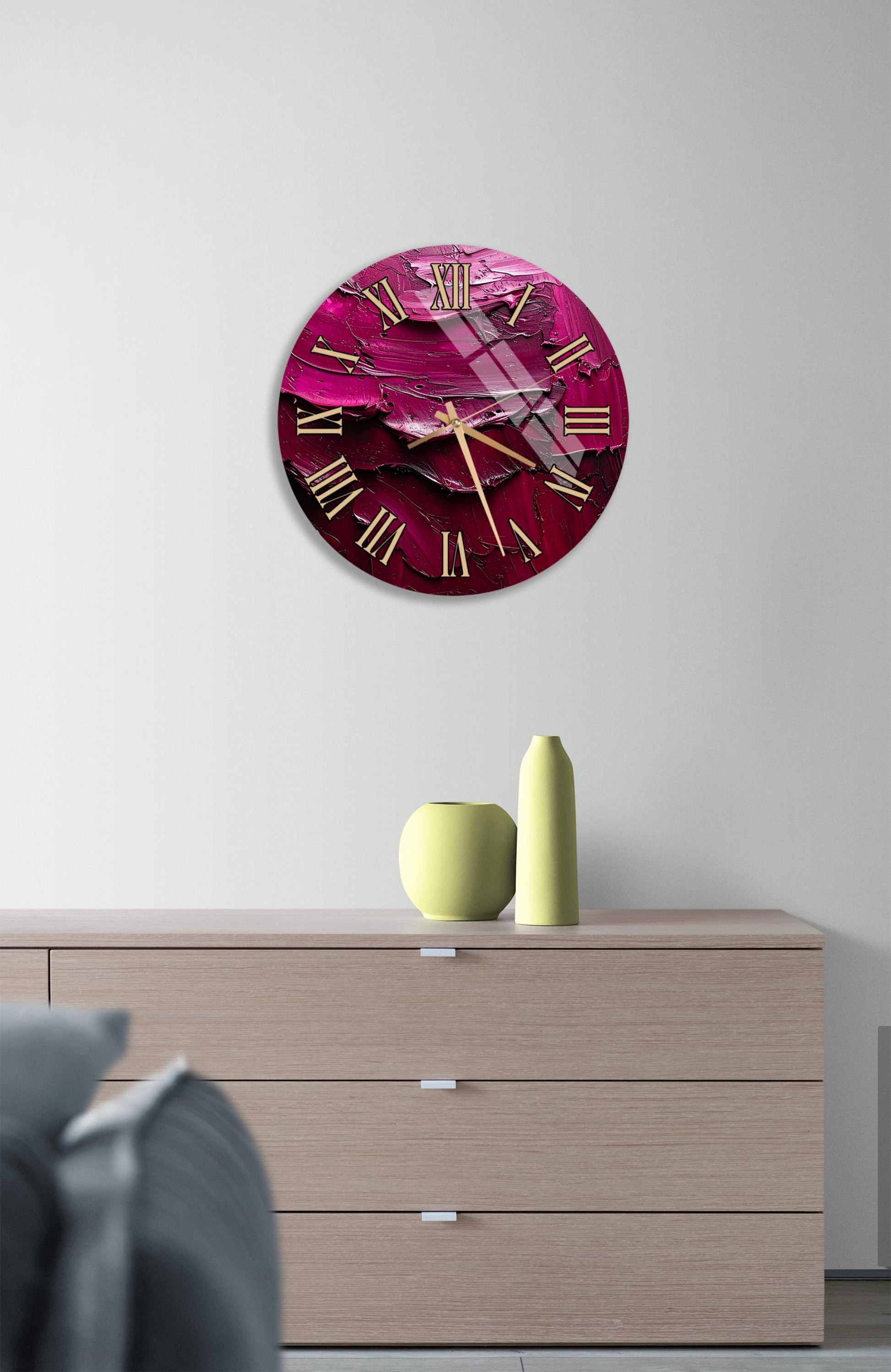 a large, round clock with a pink and purple color scheme, mounted on a wall above a wooden dresser. The dresser has a vase and a small sculpture on top of it.