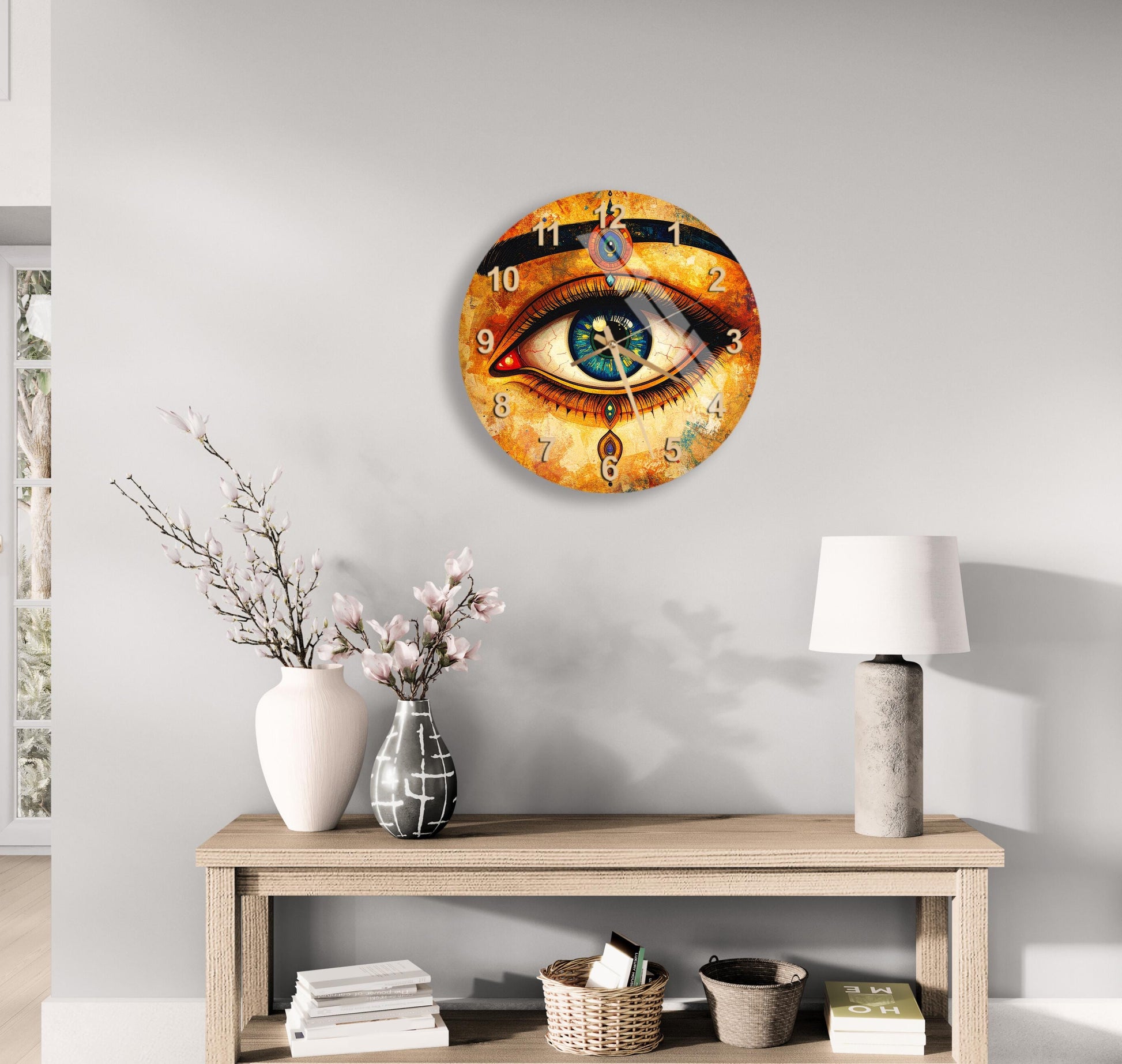 a wall clock with an eye design, a wooden table with a lamp, a vase with flowers, and some books on a shelf.