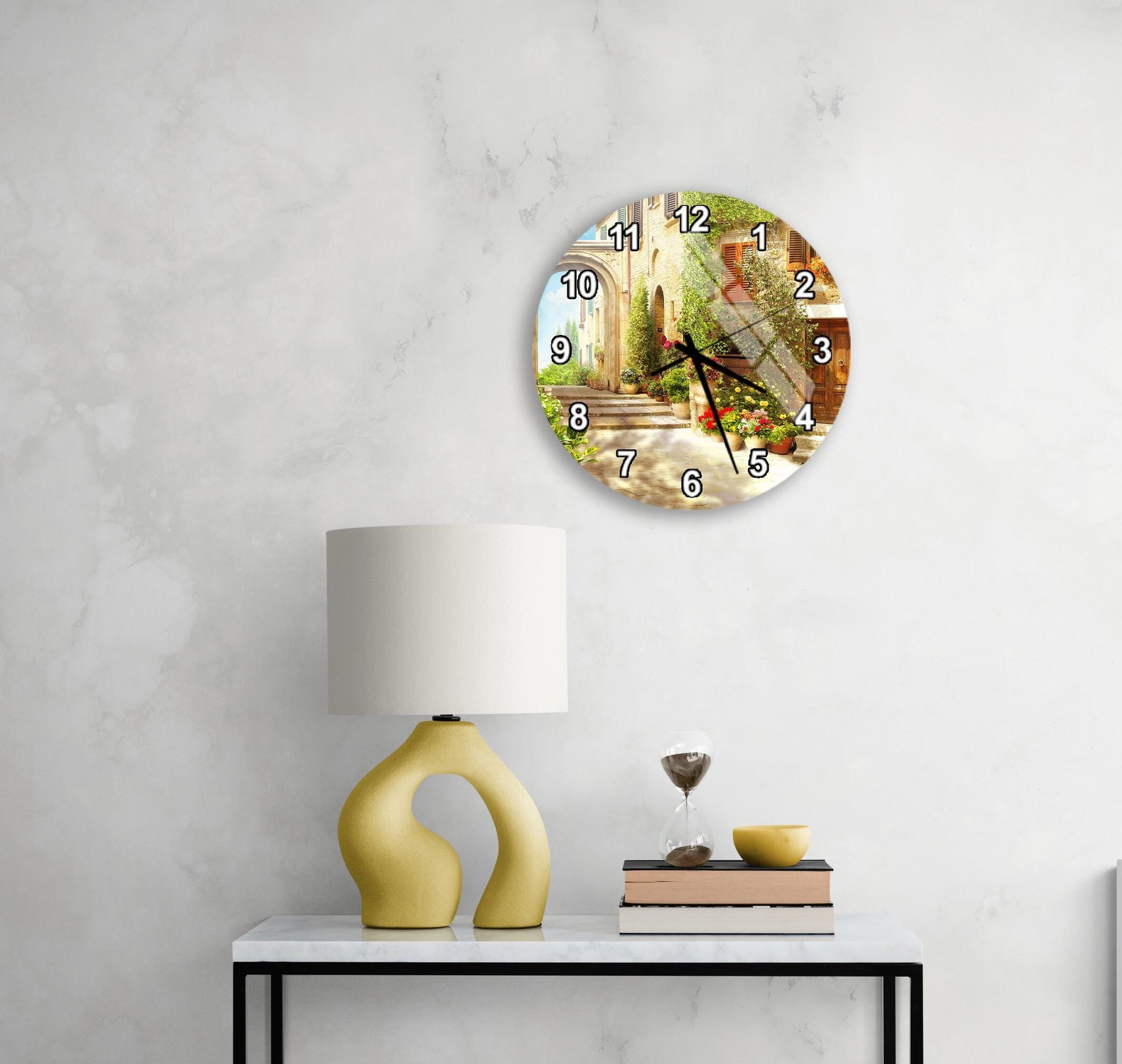 a wall clock with a scenic outdoor view, a yellow lamp on a table, and a stack of books on a table.
