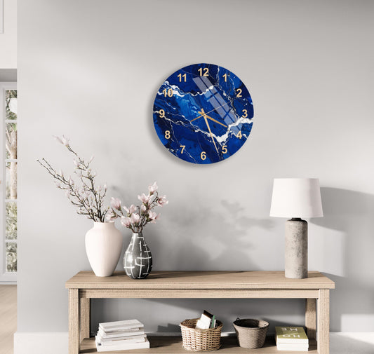 a modern living room with a large blue and white marble clock hanging on the wall above a wooden table. The table has a lamp, a vase with flowers, and some books on it. The overall decor has a minimalist and elegant style.