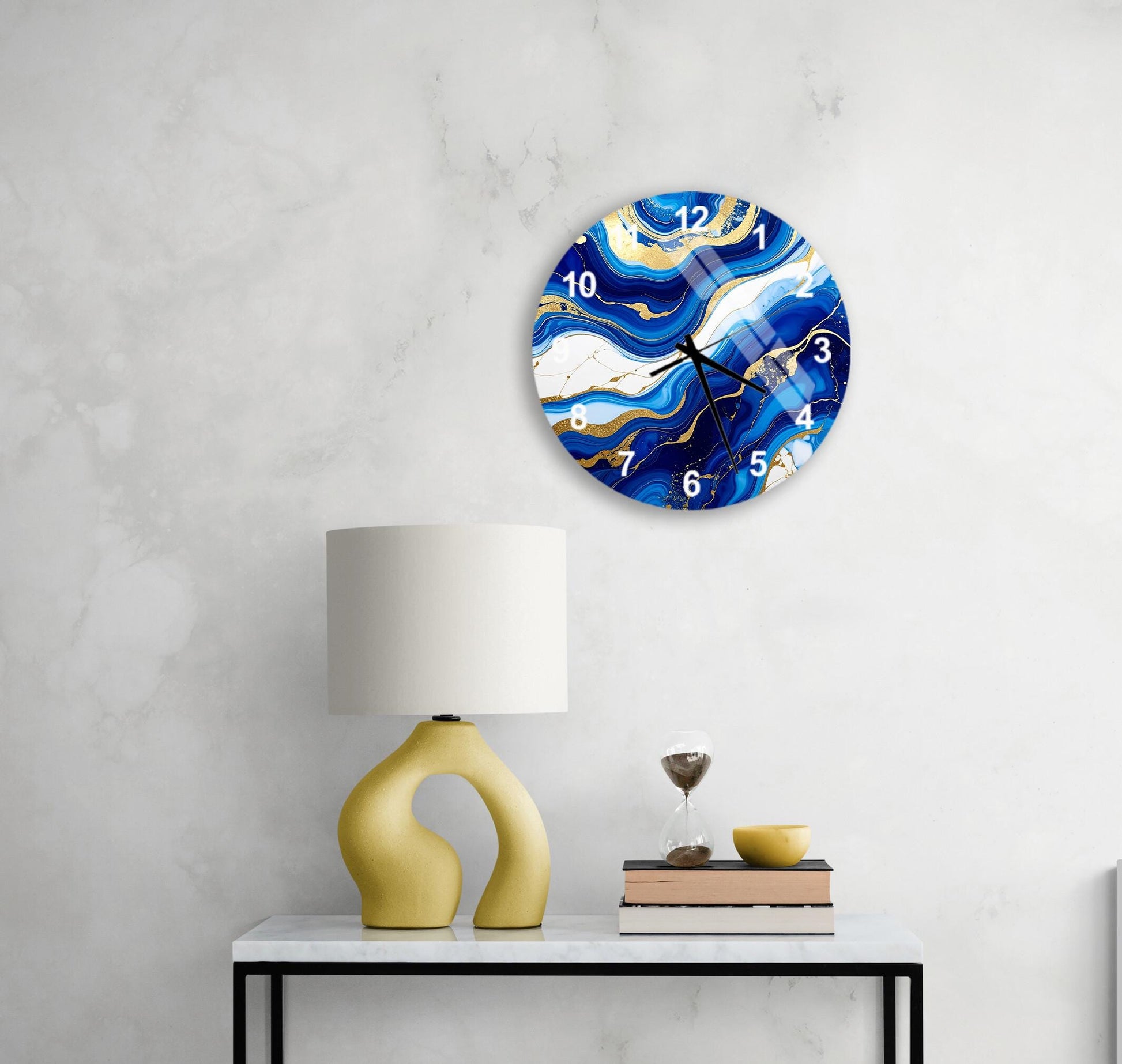 a modern wall clock with a blue and white marble design, mounted on a white wall above a white table with a yellow lamp and a stack of books.