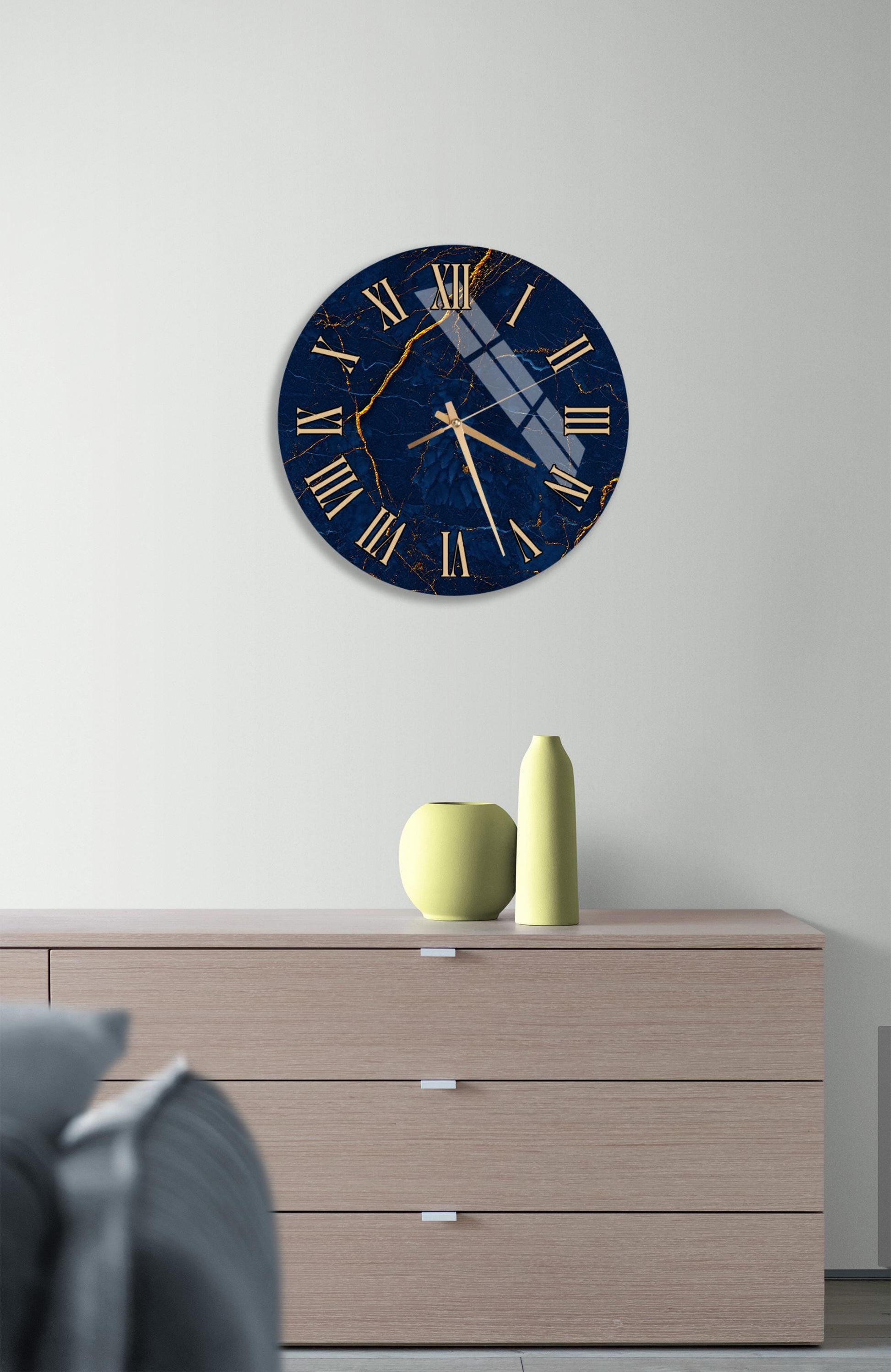 a wall clock with a blue and gold design, mounted on a white wall above a wooden dresser. The dresser has a vase and a small yellow object on top of it.