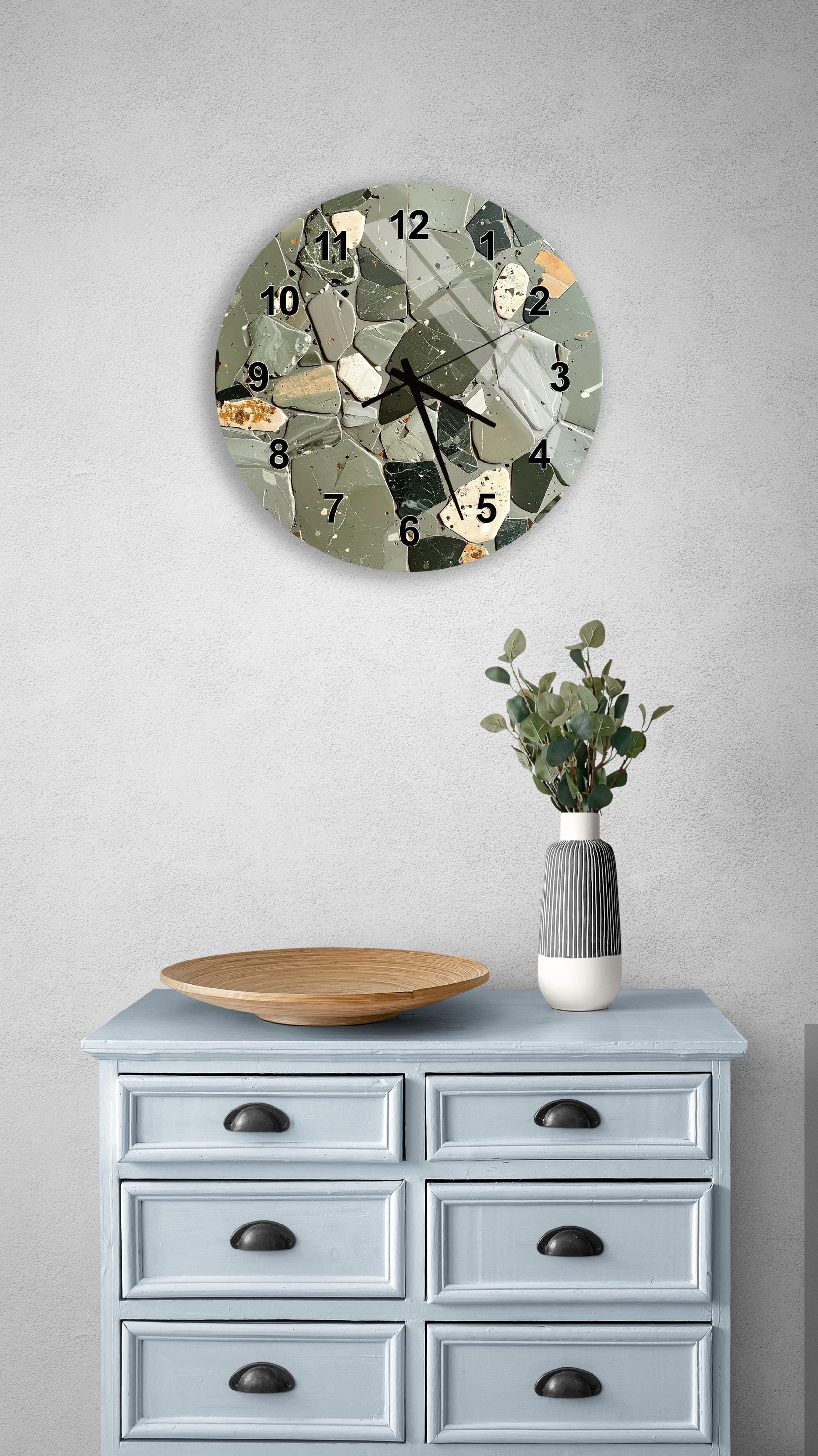 a wall clock with a mosaic design, a wooden bowl, and a vase with greenery on a light blue dresser.