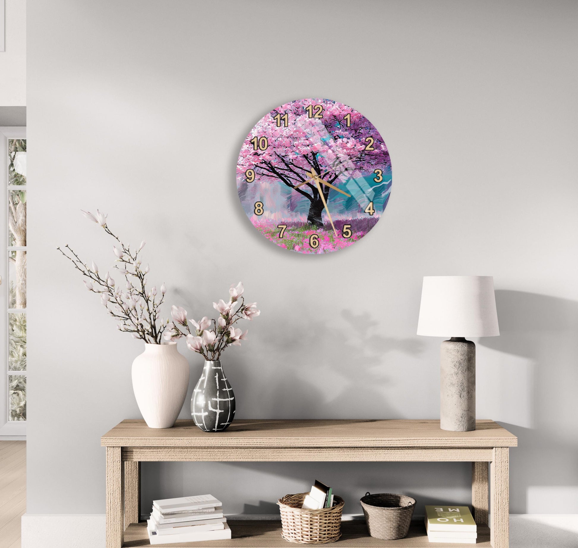 a wall clock with a pink tree design, a wooden table with a lamp, a vase with flowers, and some books on a shelf.