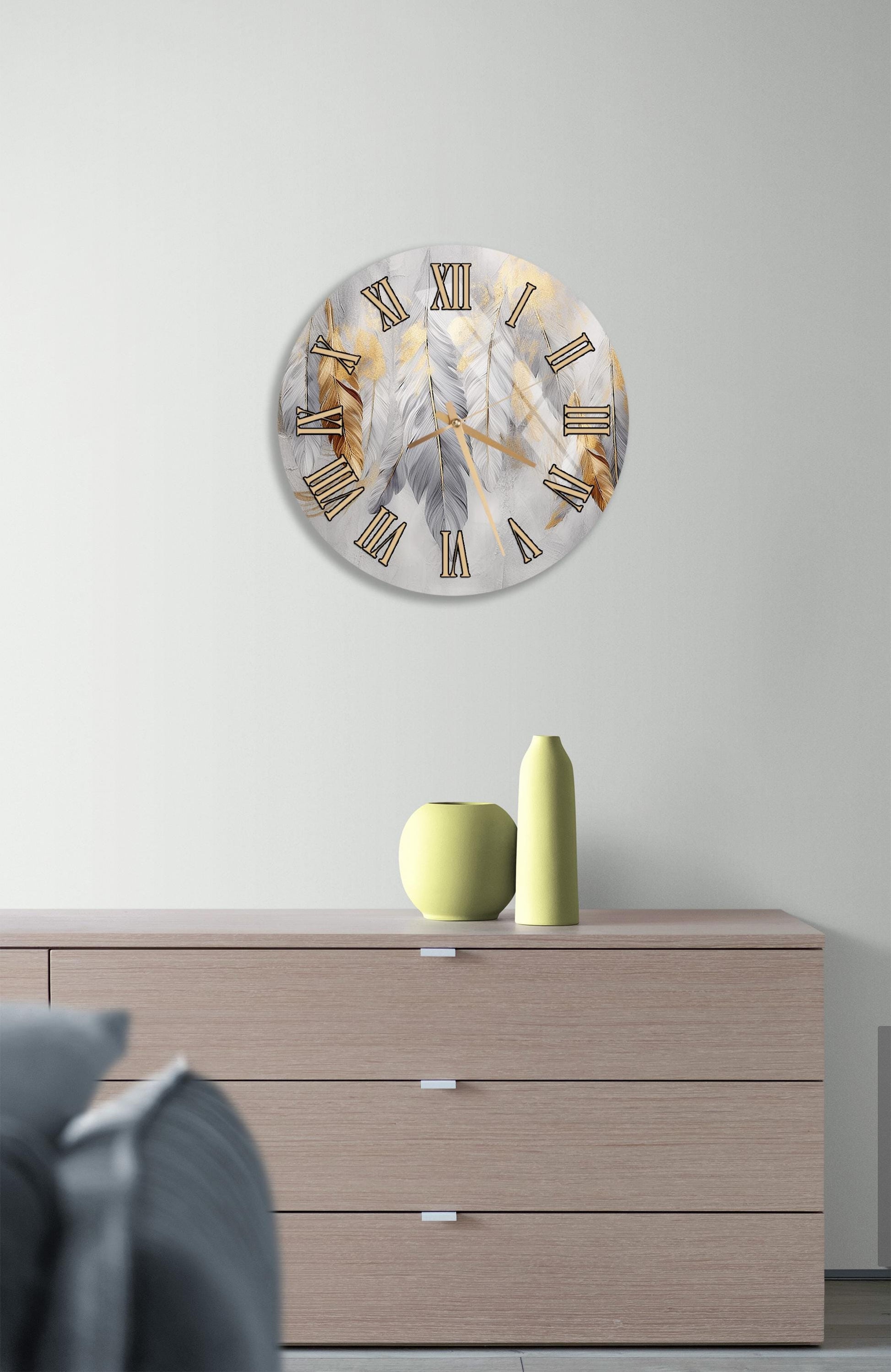 a modern living room with a round clock featuring a feather design on the wall above a wooden dresser. The dresser has three drawers and contains a yellow vase and a small green pot.