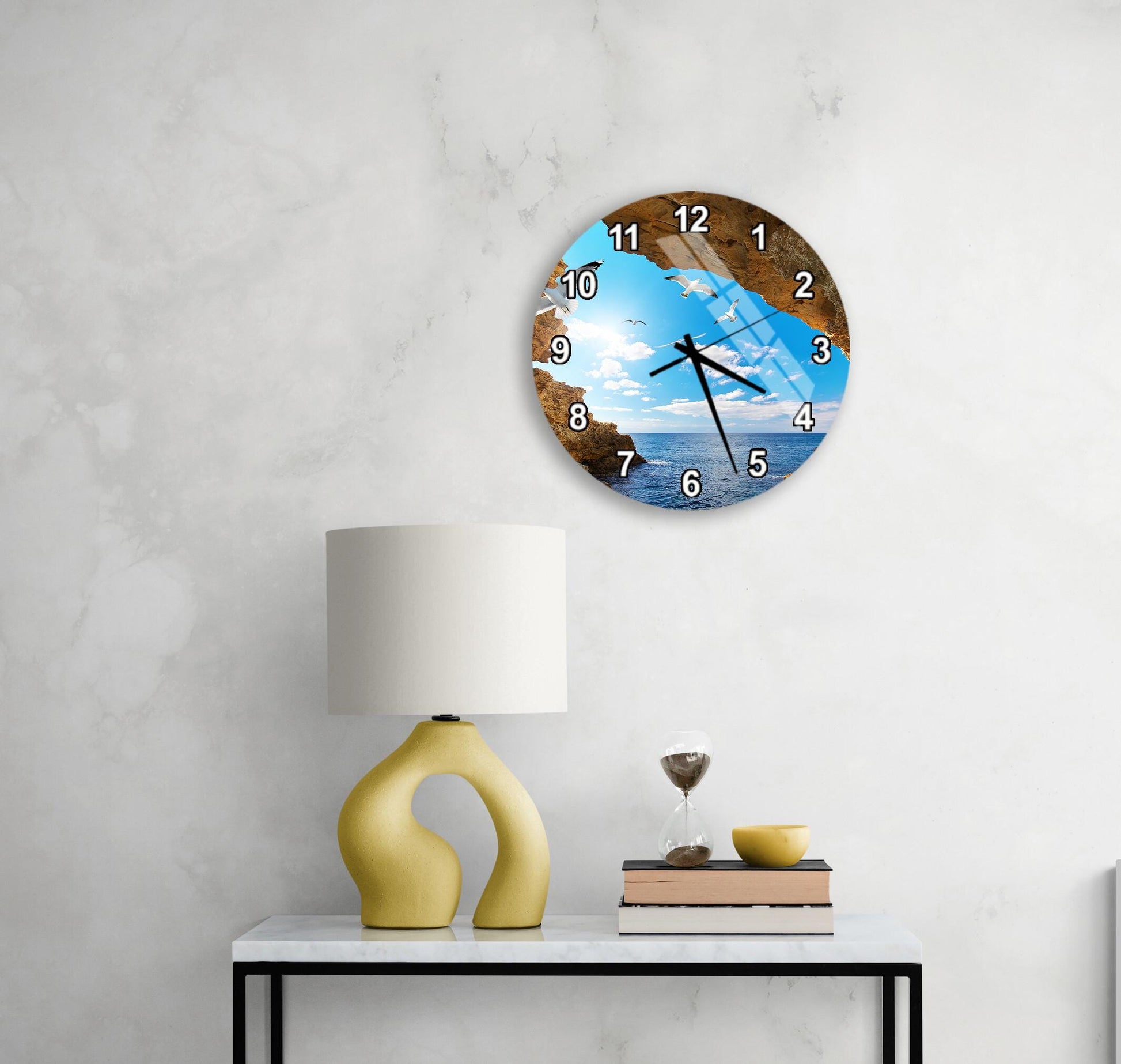 a wall clock with a beach scene on its face, mounted on a wall above a table with a lamp and a glass of water.