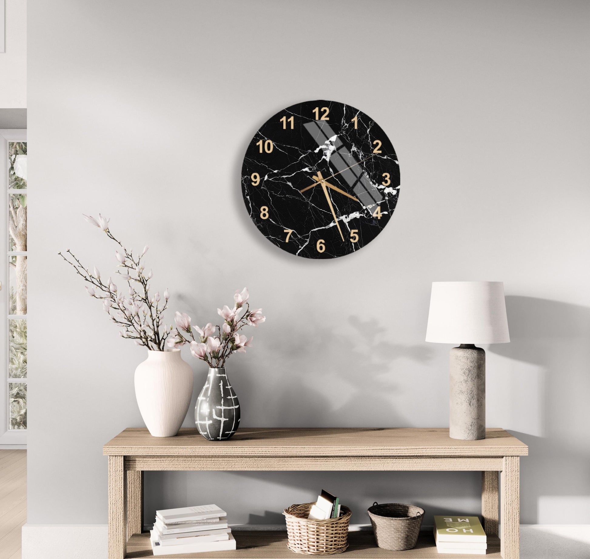 a modern black and white clock with gold accents, mounted on a wall above a wooden table with a lamp and decorative items.