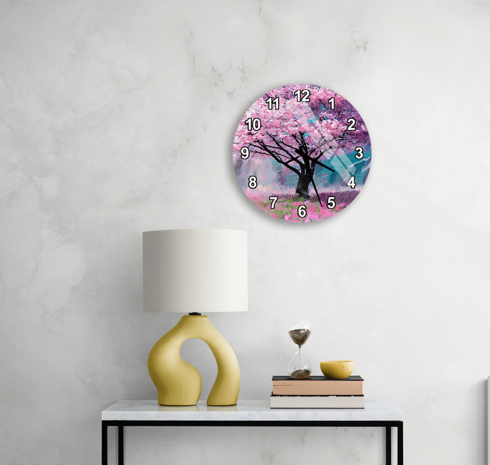 a round clock with a pink tree design on the wall above a white table with a yellow lamp and a stack of books.