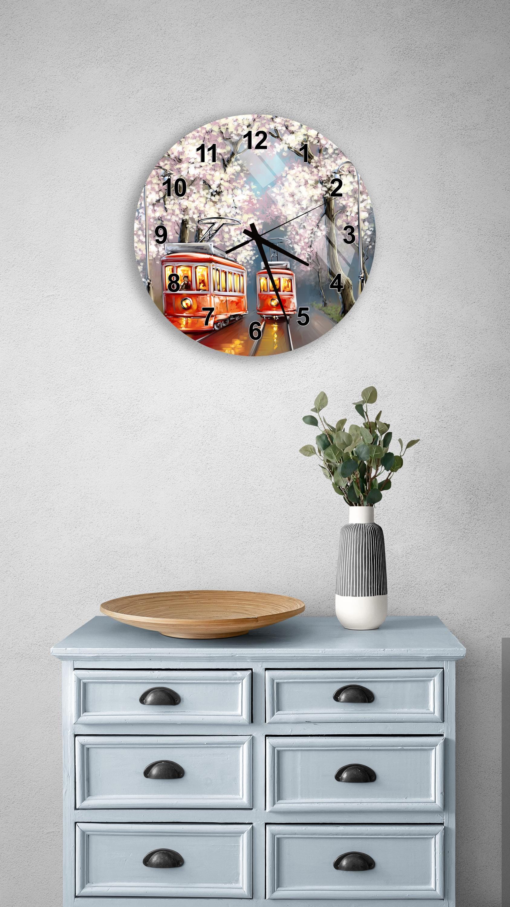 a wall clock with a trolley car design, mounted on a wall above a light blue dresser with six drawers. The dresser also has a vase of greenery and a wooden bowl on top of it.