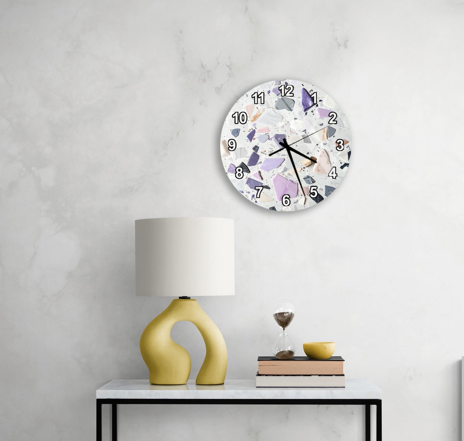 a white wall with a round clock displaying a colorful mosaic design, a yellow lamp with a unique curved base, a stack of books, and a small glass vase with a single flower.