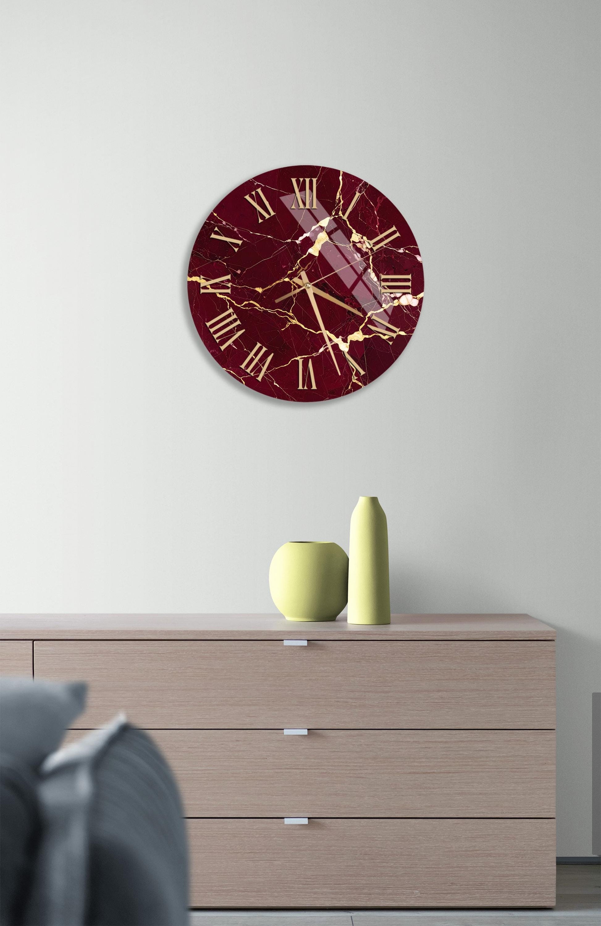 a modern interior design with a large burgundy-colored clock with gold accents hanging on a white wall above a wooden dresser. The dresser has three drawers and a small yellow vase on top.