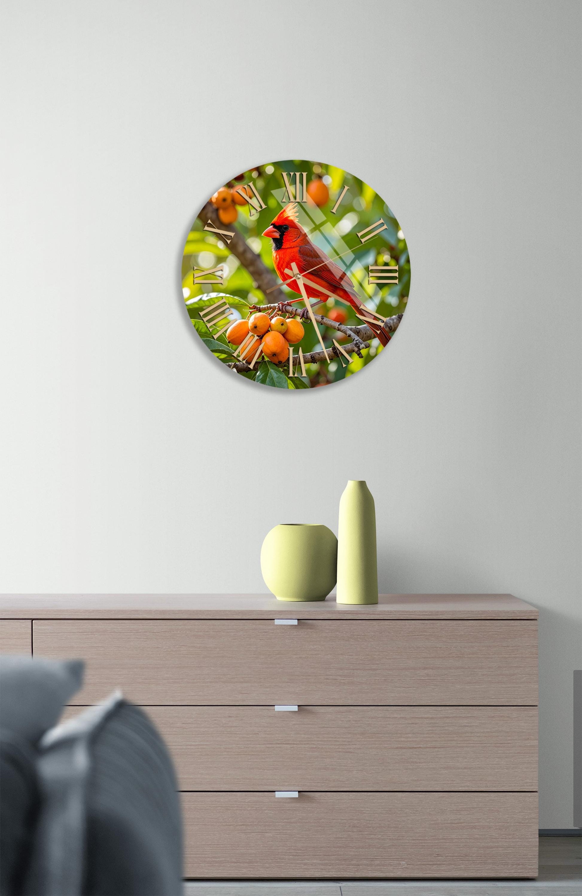 a round clock with a vibrant red cardinal perched on a branch, surrounded by orange berries, hanging on a white wall above a wooden dresser.