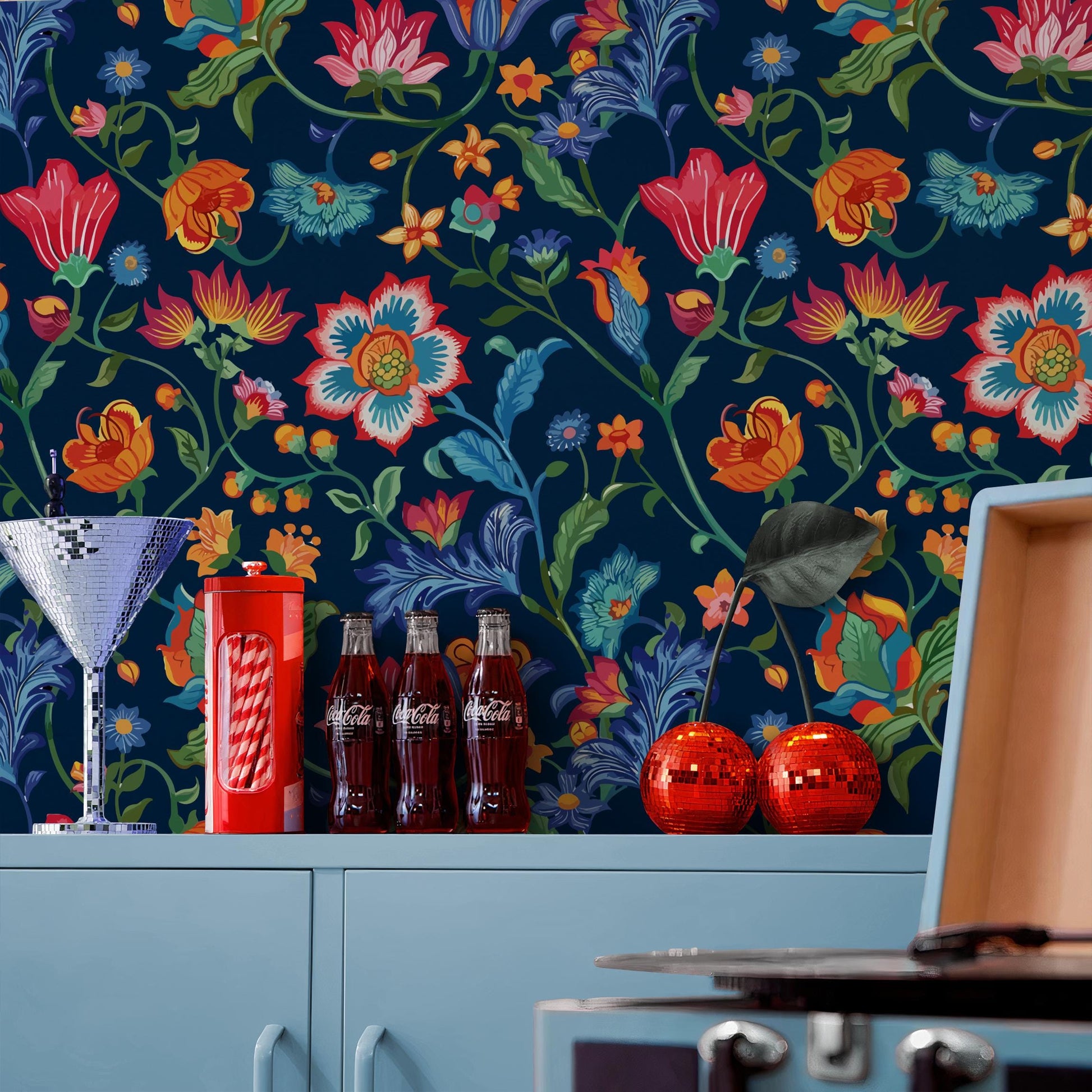 a vibrant floral wallpaper with a variety of colorful flowers and leaves, including red, orange, and yellow flowers. On a shelf below the wallpaper, there are three Coca-Cola bottles, two red balls, and a silver cocktail glass.
