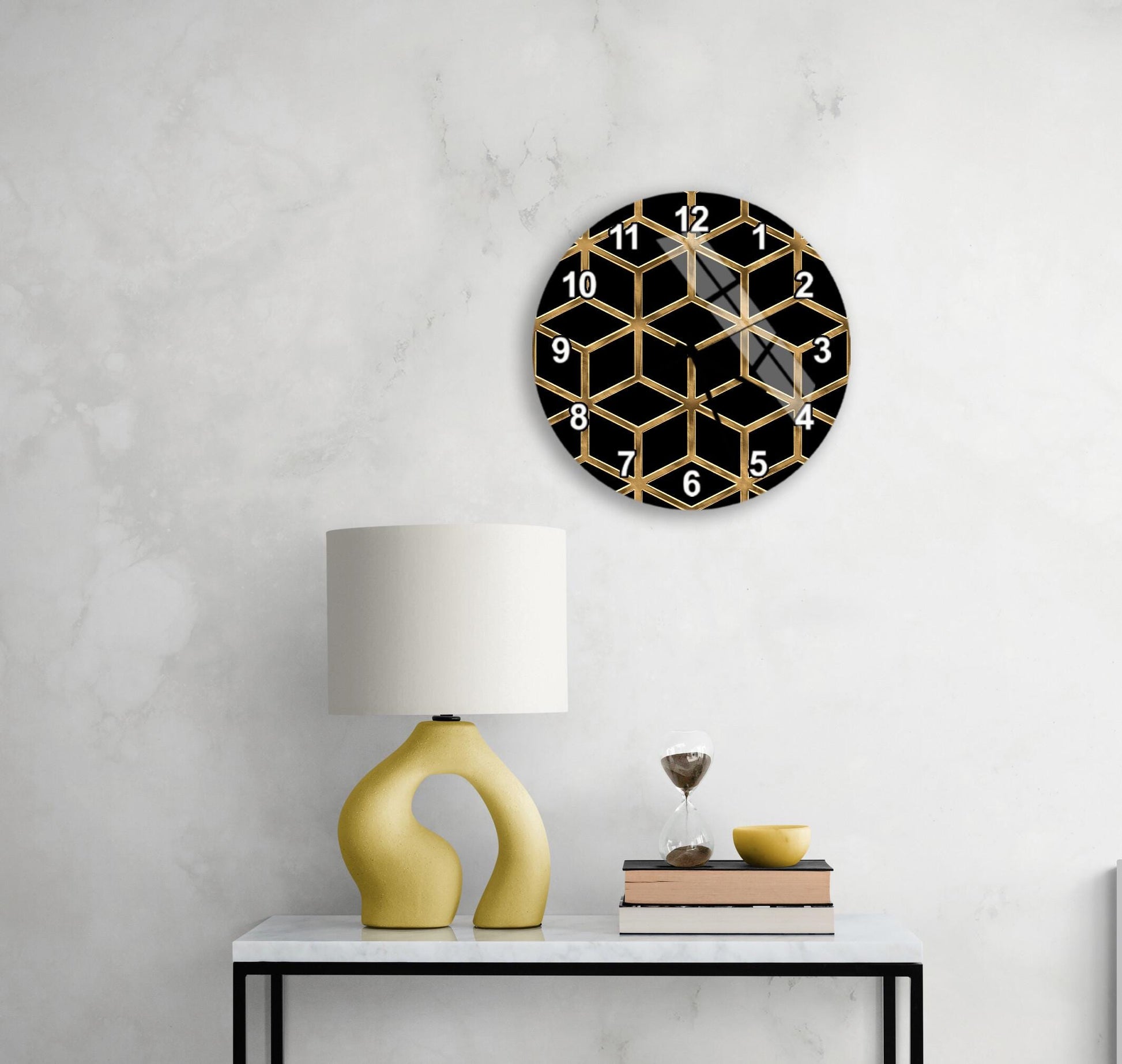 a modern black and gold geometric clock mounted on a white wall above a white table with a yellow lamp and a stack of books.