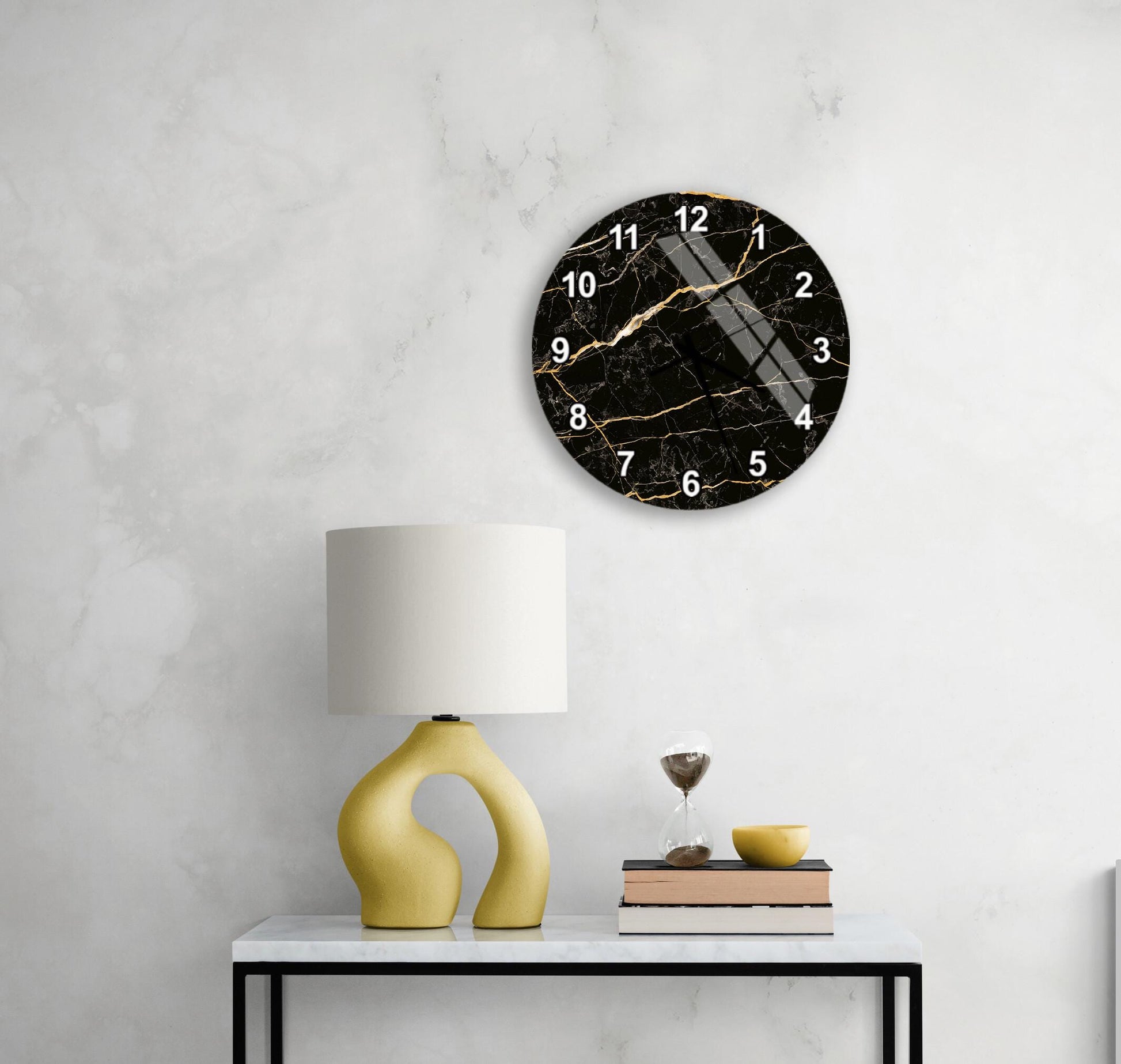 a black marble clock with gold accents, mounted on a white wall above a white table with a yellow lamp and a stack of books.