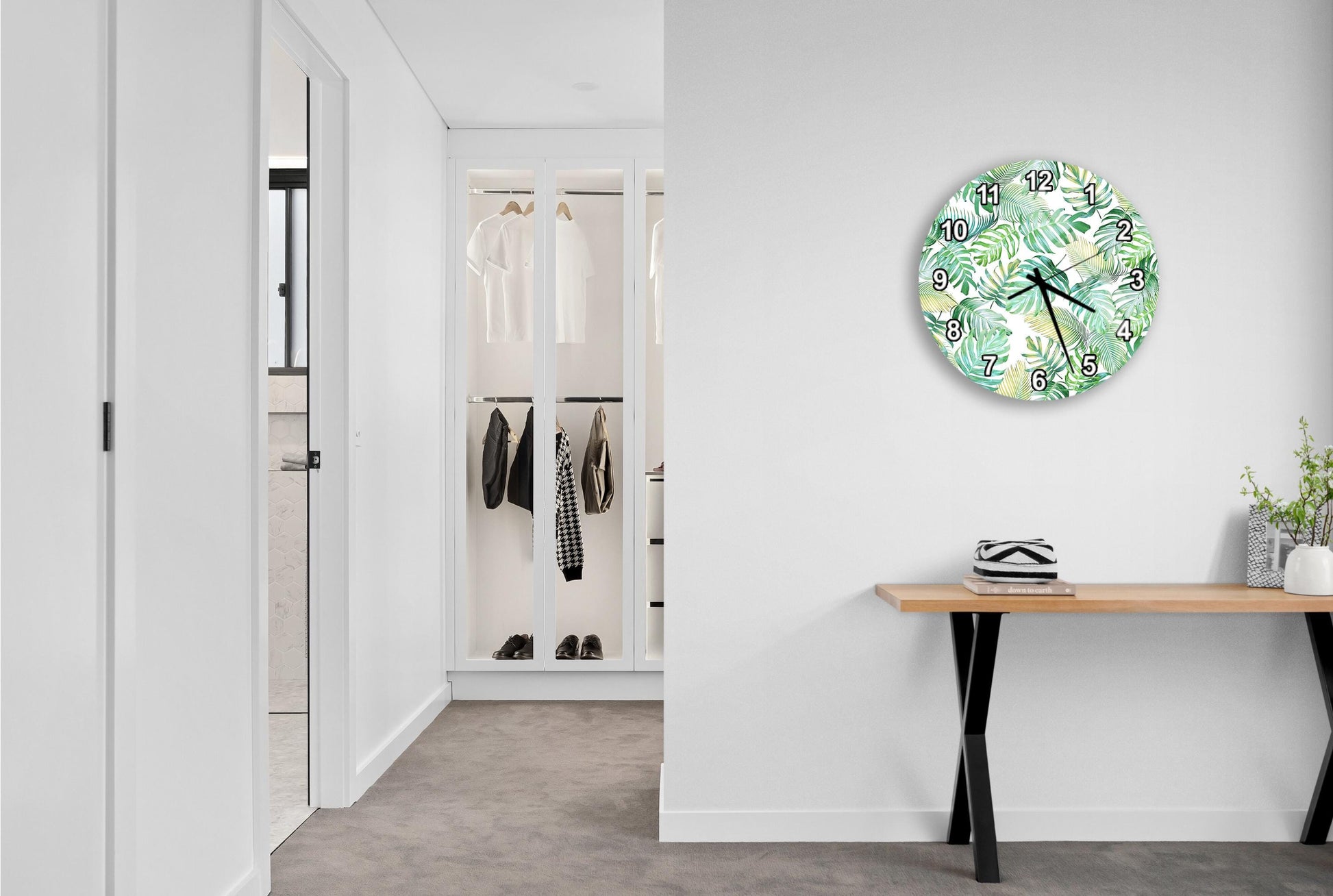 a modern, minimalist hallway with a large, round wall clock featuring a tropical leaf pattern. The hallway is lined with white walls and features a wooden console table with a plant on top.