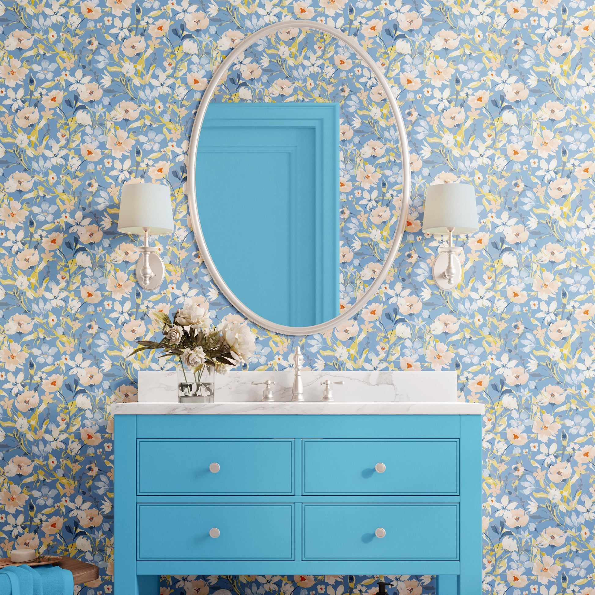 a bathroom with a blue vanity and a mirror above it. The vanity has a white countertop and is accompanied by two white lamps on either side. The wall behind the vanity is covered in a floral wallpaper, and there is a vase of flowers on the vanity.