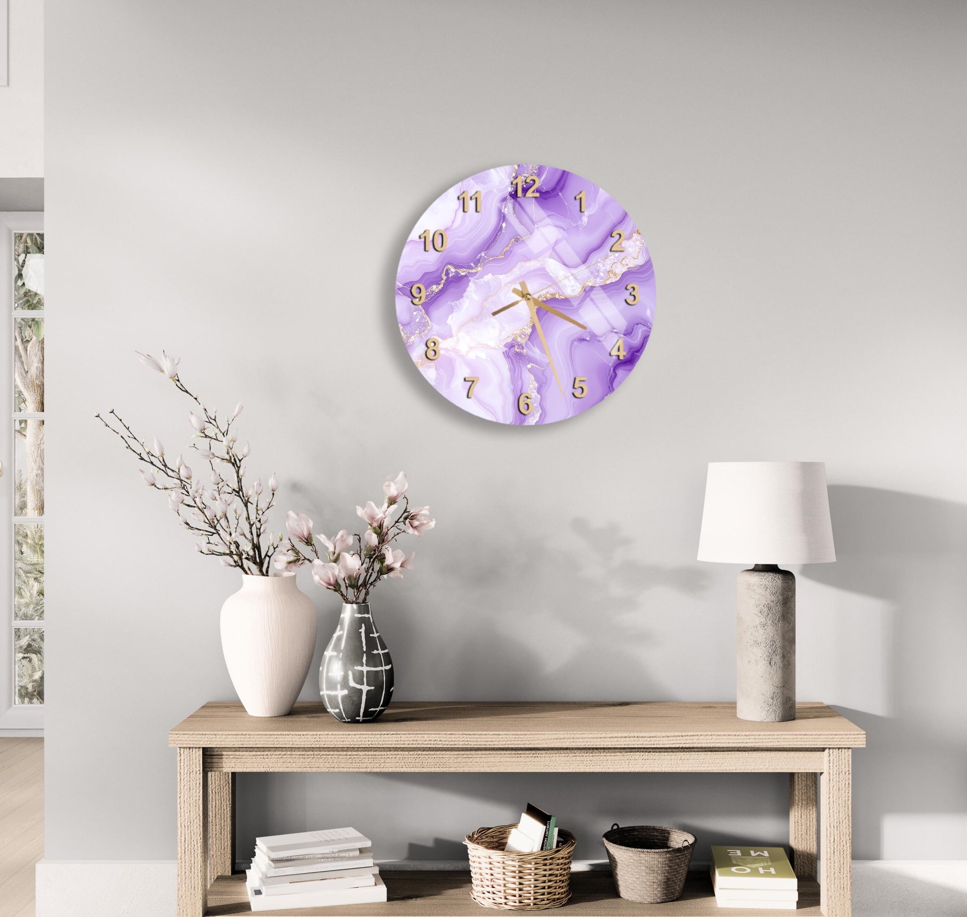 a modern and minimalist living room with a round clock featuring a purple marble design on the wall. The clock is mounted above a wooden table with a lamp and a vase of flowers. The table also holds a stack of books and a basket.