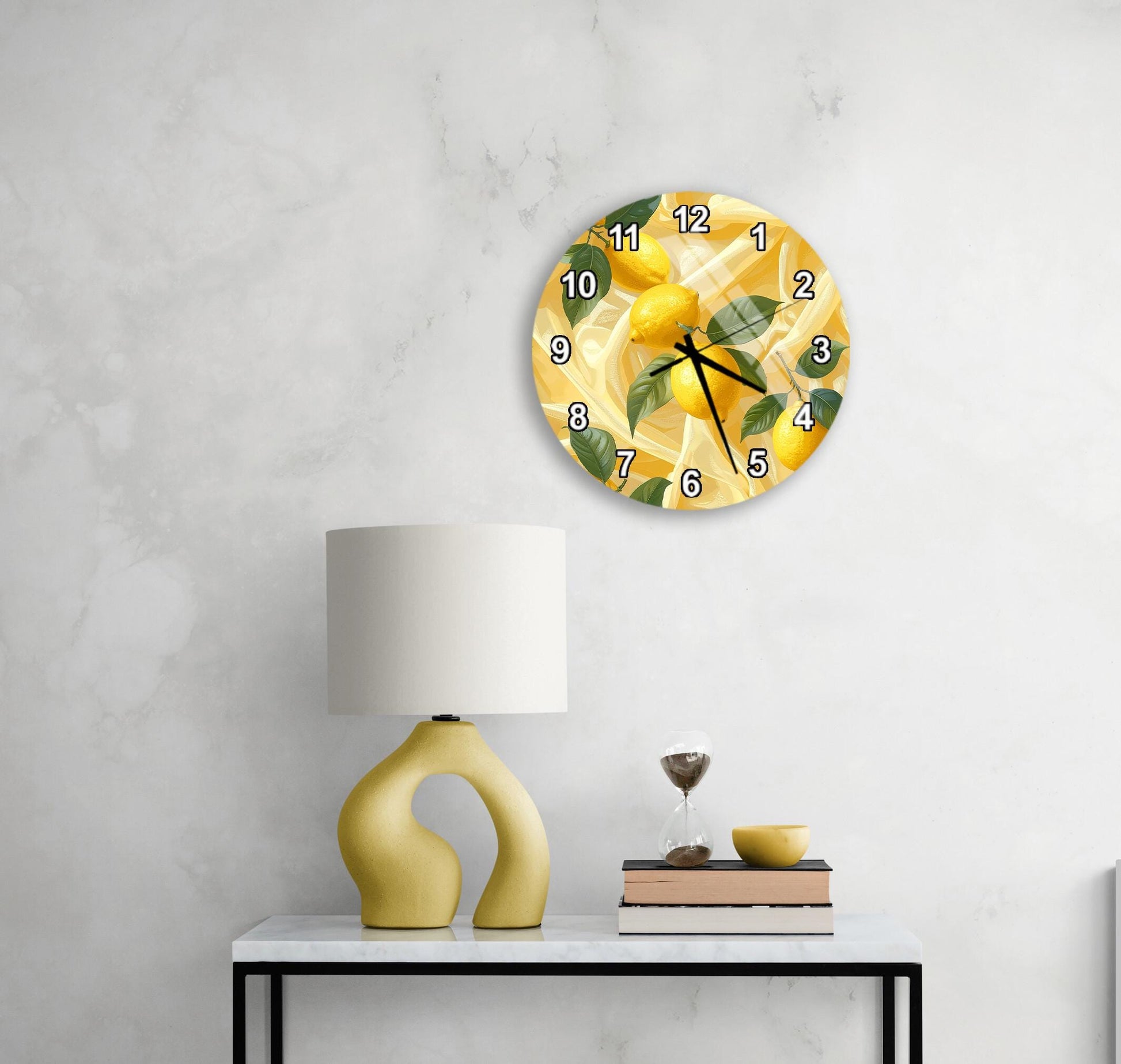 a white wall with a round clock displaying a lemon design, a yellow lamp, a stack of books, and a glass of wine on a white table.