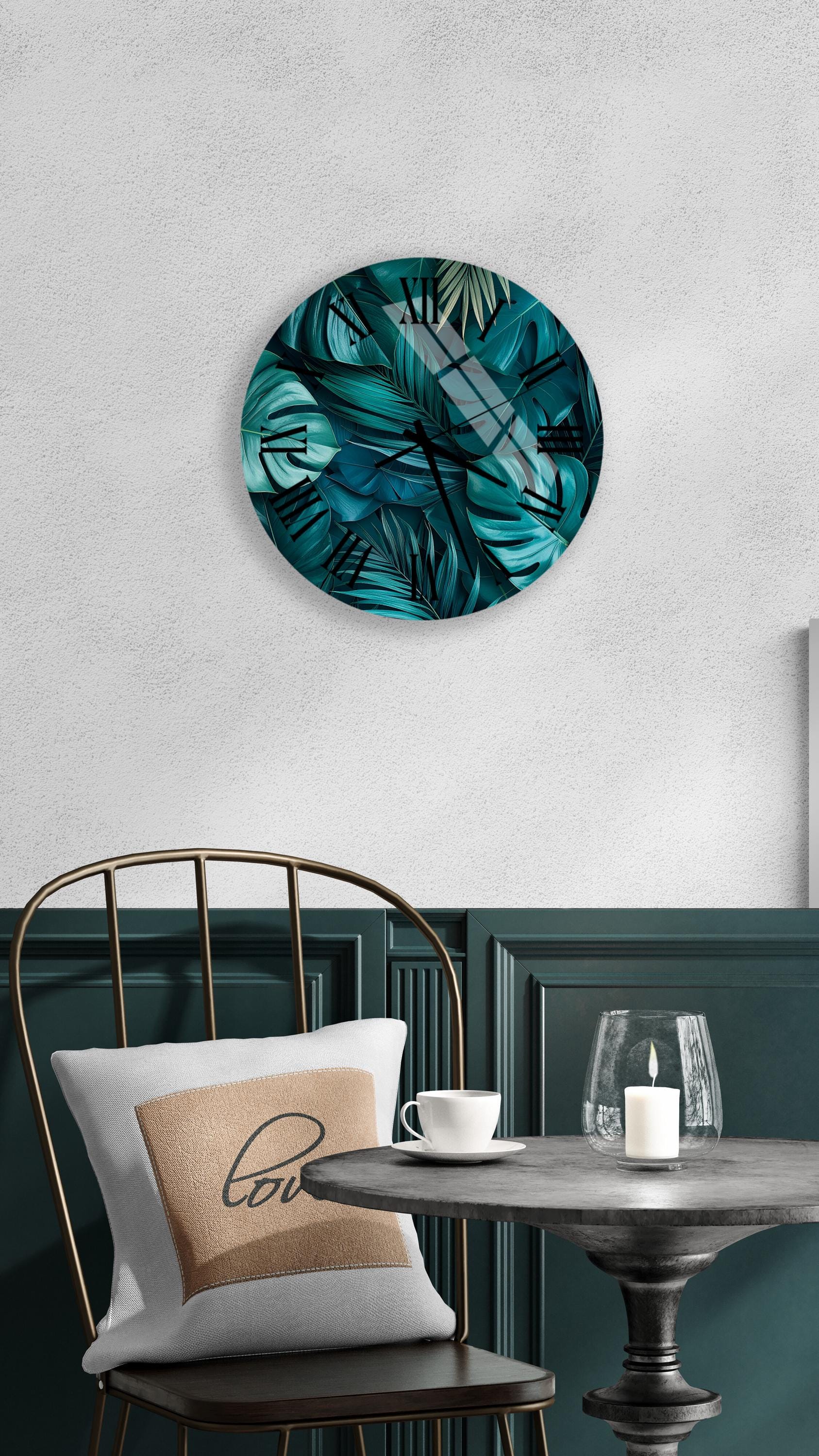 a modern and stylish interior design with a round wall-mounted artwork depicting a tropical plant pattern in shades of blue and green. The artwork is mounted above a black metal chair with a white pillow featuring a black cursive "love" design. In front of the chair, there is a small table with a glass candle holder and a white teacup.
