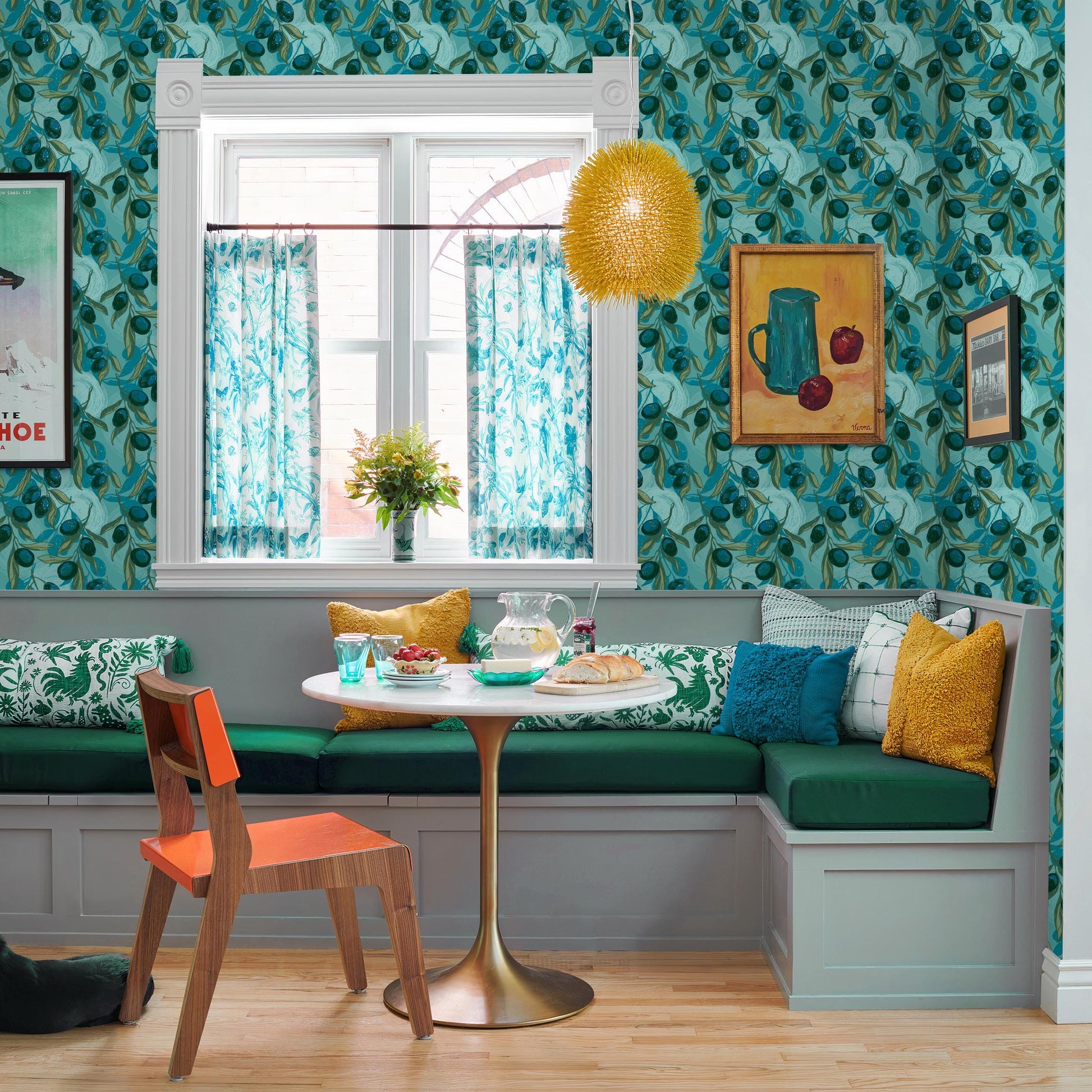 a cozy dining area with a round table, green cushions, and a wooden bench. A yellow pendant light hangs above the table, and a painting of a vase with flowers is displayed on the wall. The room has a vibrant and eclectic decor, with a mix of patterns, colors, and textures.