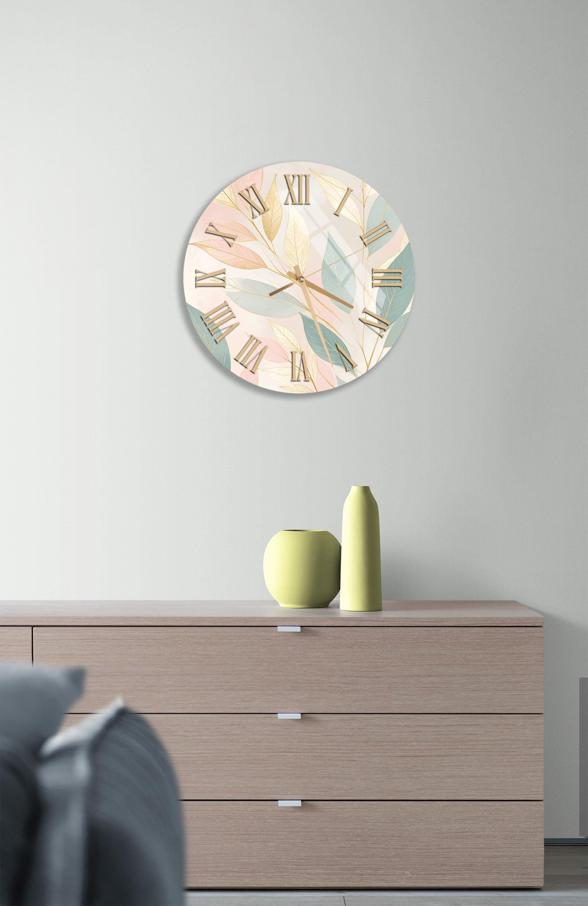 a wall clock with a leafy design, a wooden dresser with a vase and a small plant, and a gray couch in the background.