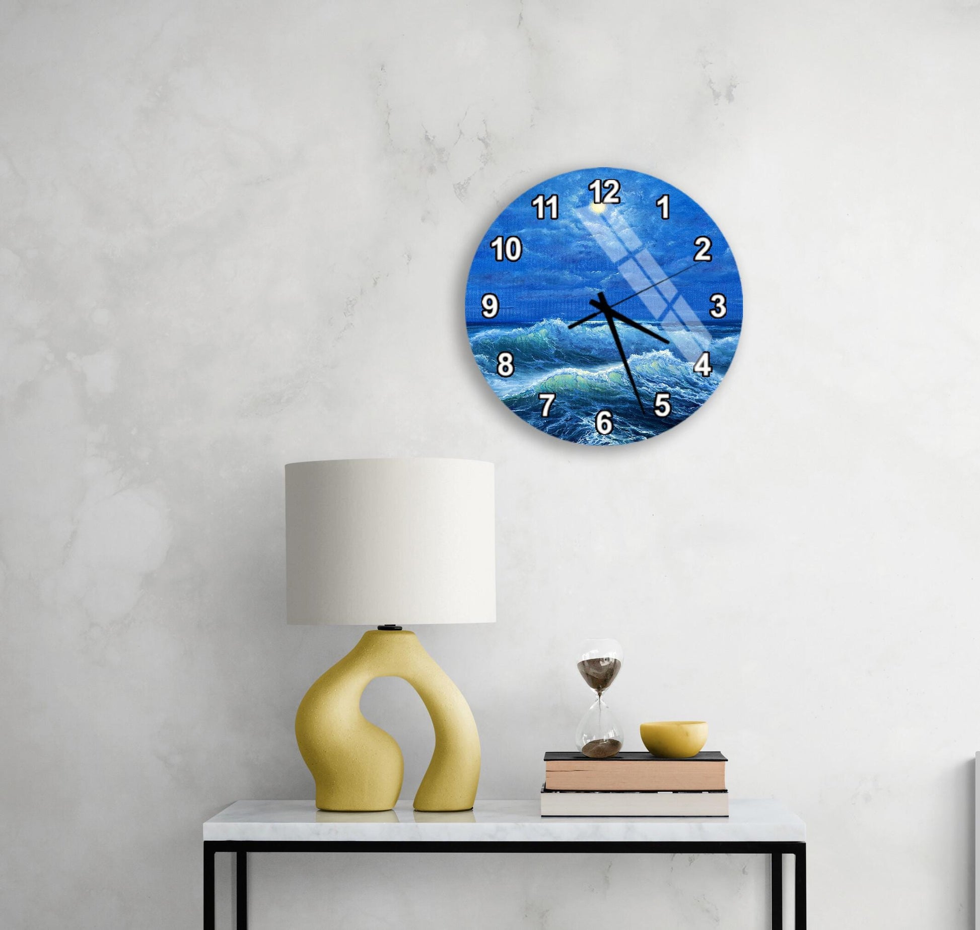 a wall clock with a blue ocean scene, a yellow lamp, a stack of books, and a glass of water on a white table.