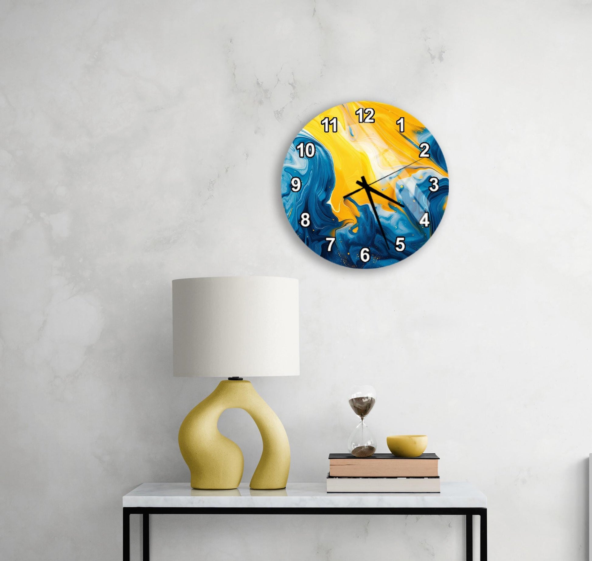 a modern wall clock with a vibrant blue and yellow design, hanging on a white wall above a white table with a yellow lamp and a stack of books.