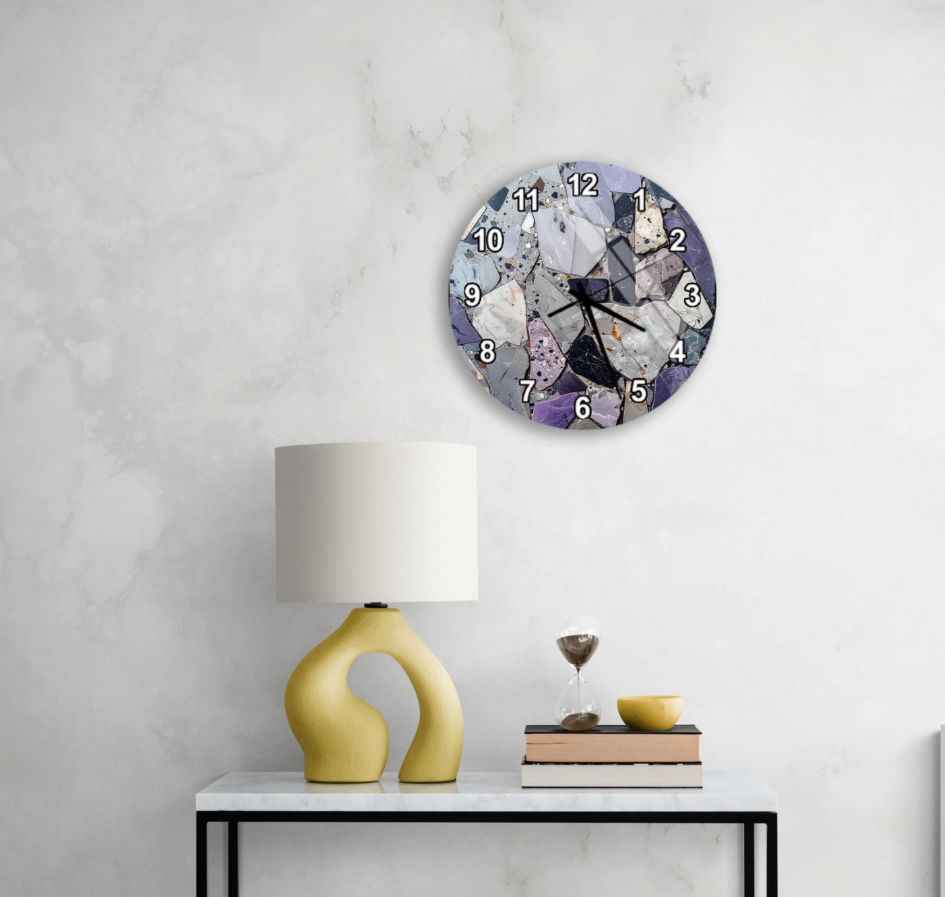 a modern clock with a unique design, mounted on a wall above a white table with a yellow lamp and a stack of books.